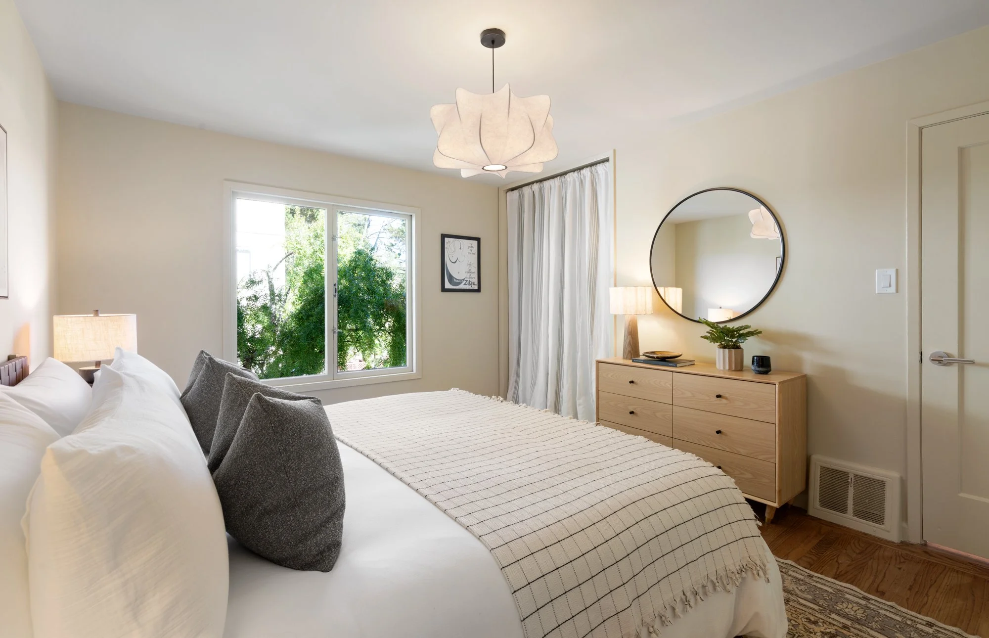 A cozy bedroom with a large bed covered in white bedding and gray throw pillows, a wooden dresser with a mirror, a table lamp, and a window with white curtains. There is a green tree visible outside the window.