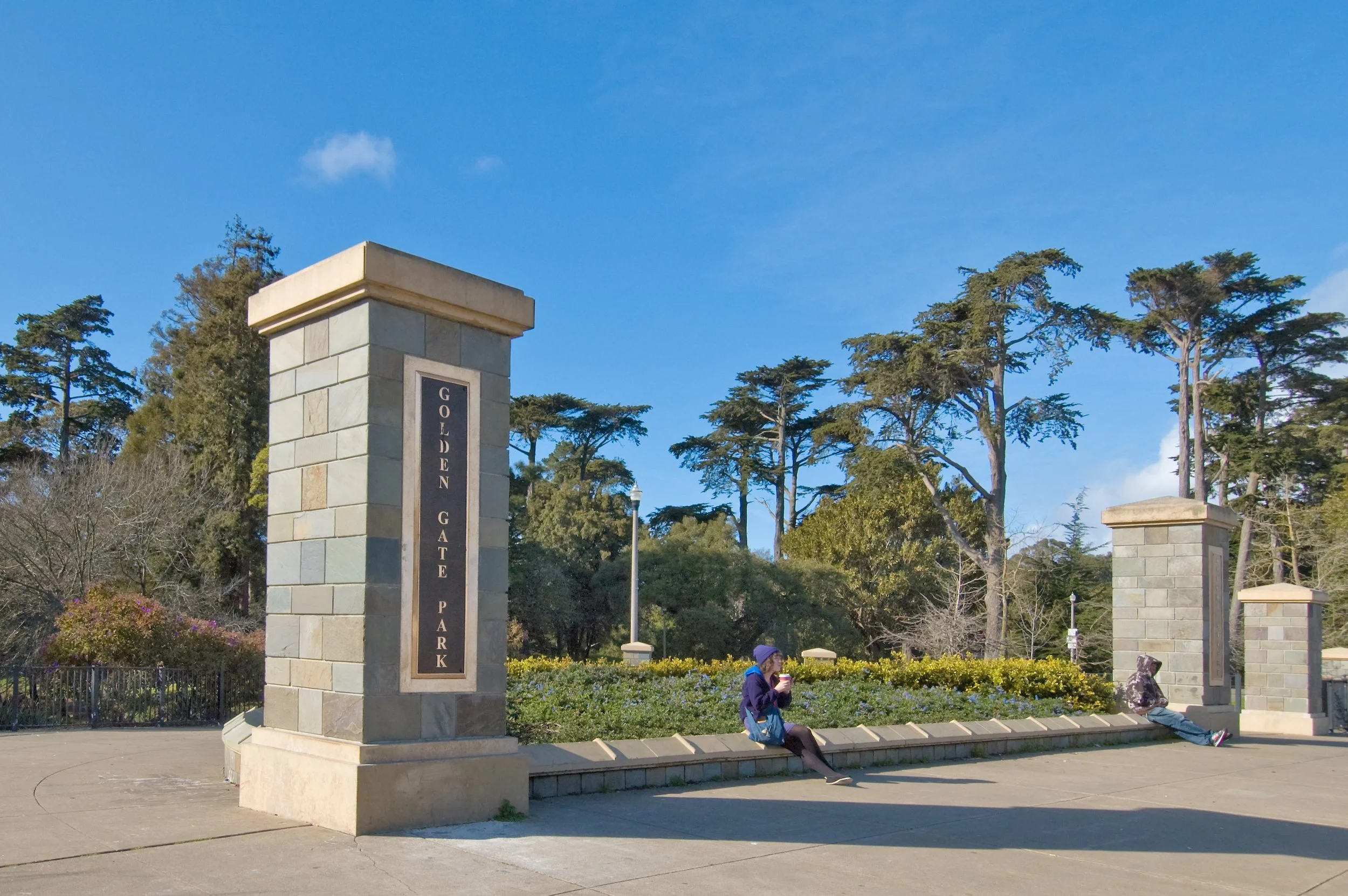 A park entrance with stone columns and a bench, trees in the background, and a woman sitting on the bench holding a drink wearing a hat.