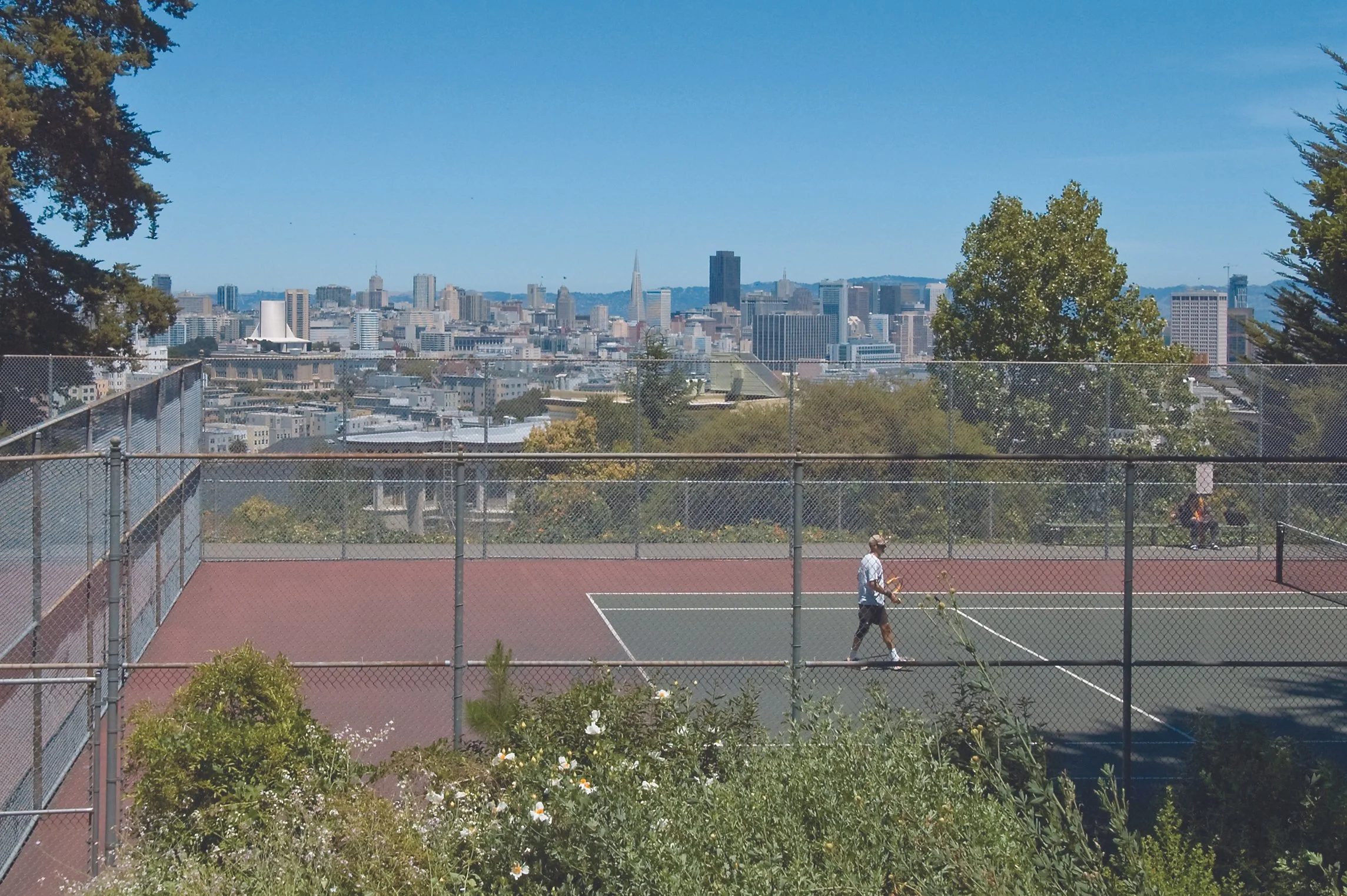 A city skyline viewed from a tennis court with a chain-link fence around it. One person is playing tennis on the court, and buildings can be seen in the background under a clear blue sky.