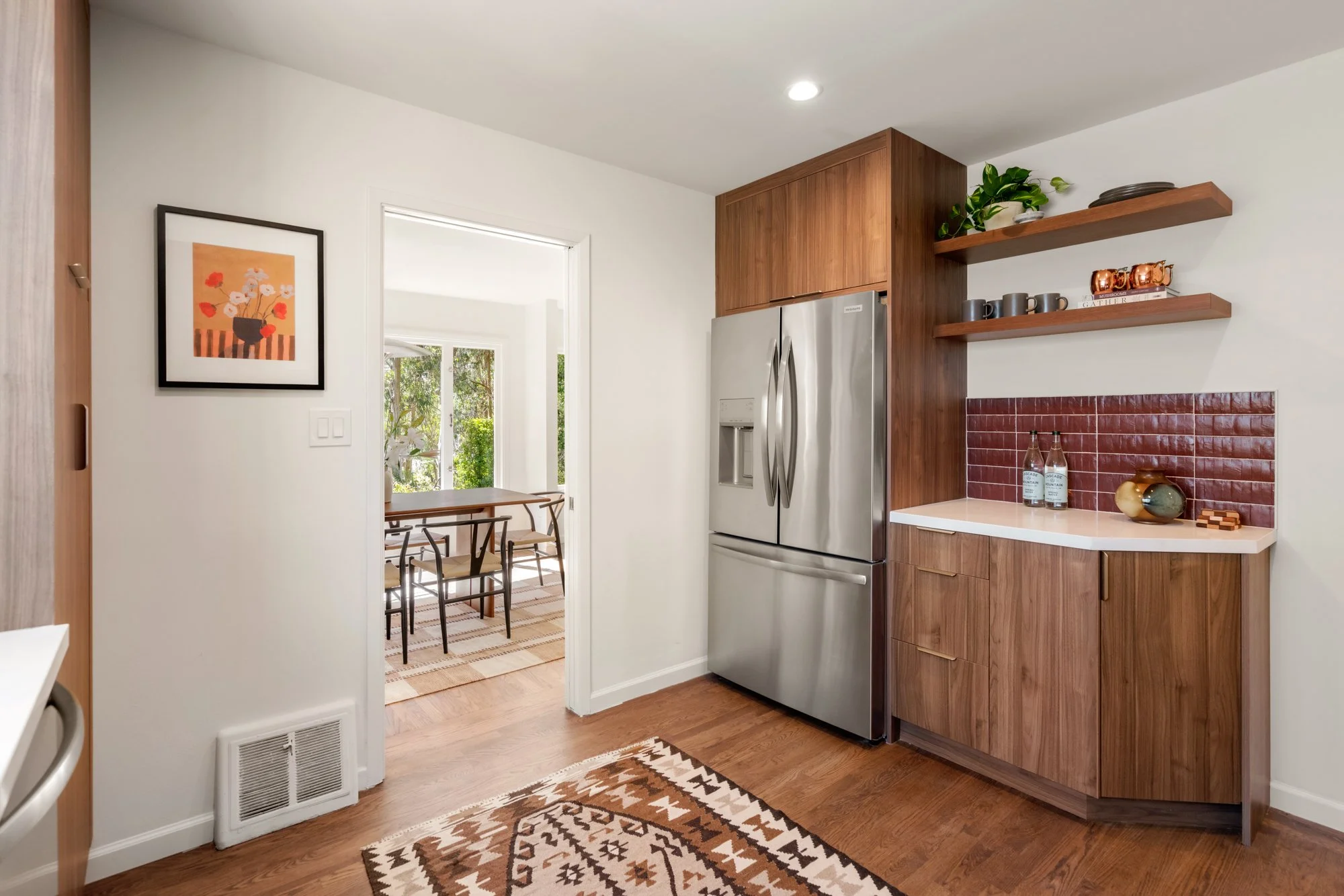 Modern kitchen corner with wooden cabinets, stainless steel refrigerator, floating shelves holding potted plant, mugs, and decorative items, red brick backsplash, and a colorful patterned rug on hardwood flooring.