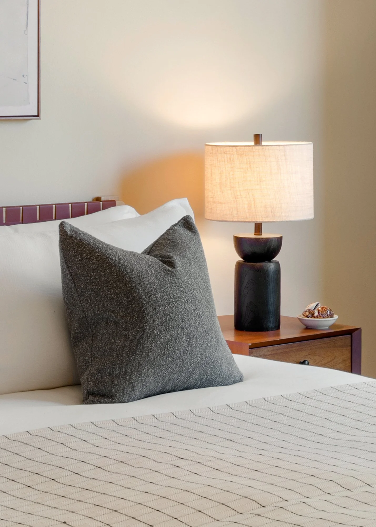 Close-up of a bed with a gray pillow and a cream-colored bedspread in a bedroom. A wooden nightstand with a black lamp with a beige shade and a small bowl of decorative seashells is next to the bed. A framed picture hangs on the beige wall above the bed.