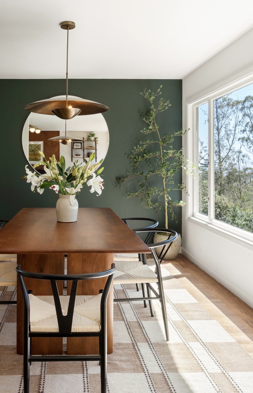A dining room with a wooden table, black chairs with woven seats, a large window, a potted tree, a mirror on a dark green wall, and a vase with white lilies and green leaves on the table. Overhead, a modern pendant light hangs.