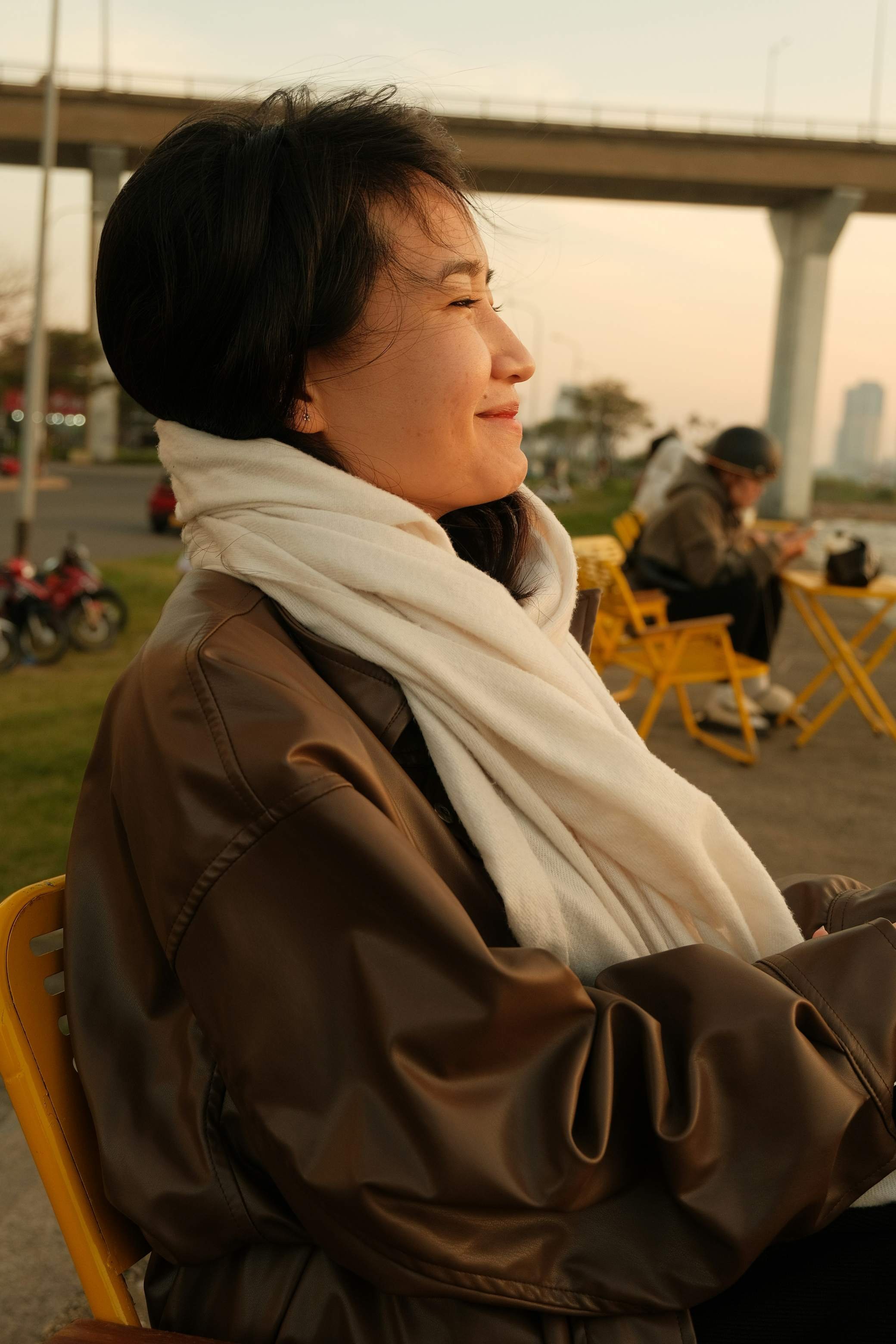 Woman in a brown leather jacket and cream scarf sitting outdoors at dusk, eyes closed and smiling softly