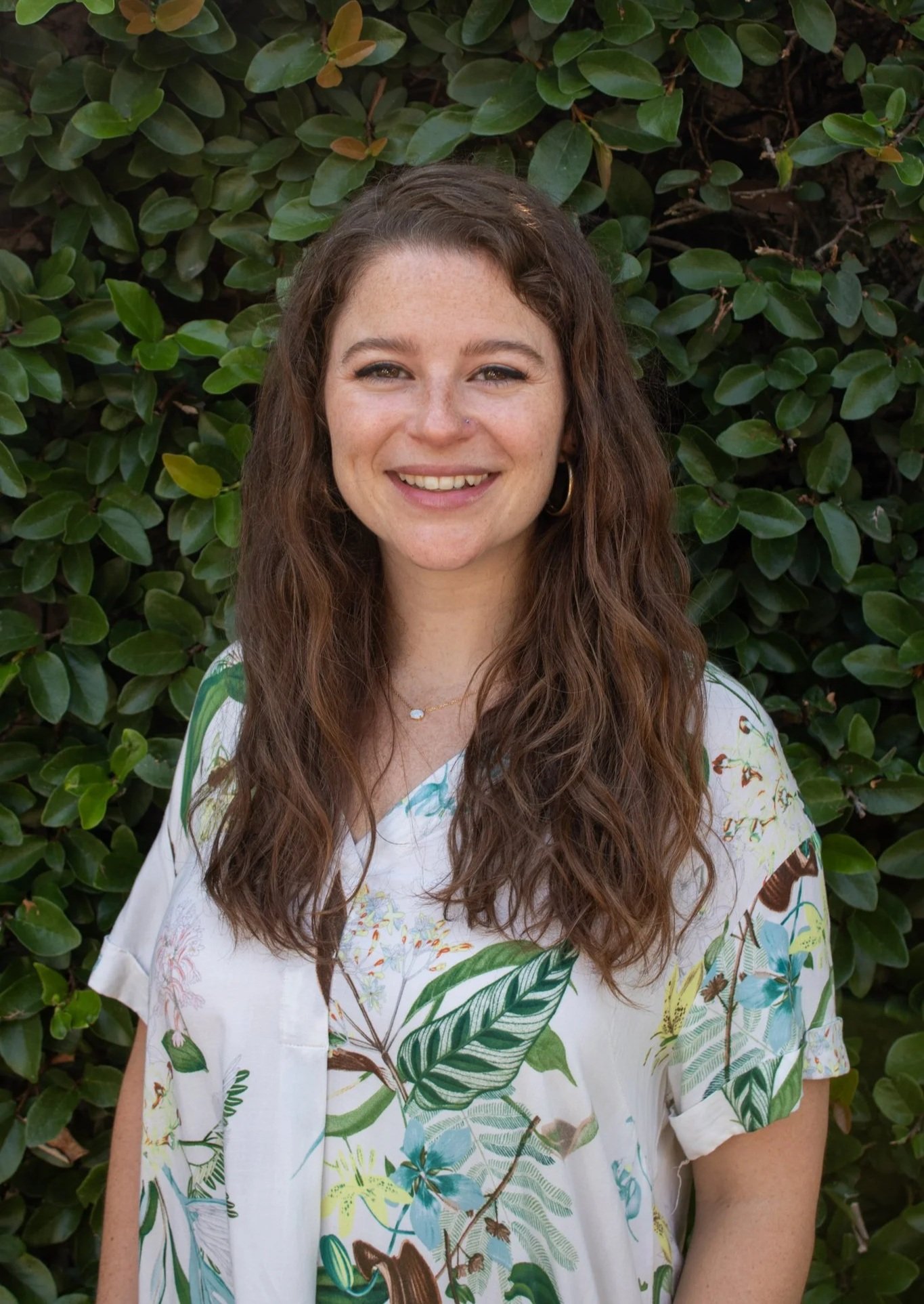 A young woman with long, wavy brown hair and light skin, smiling, standing in front of a lush green hedge, wearing a white, colorful, floral-patterned blouse.