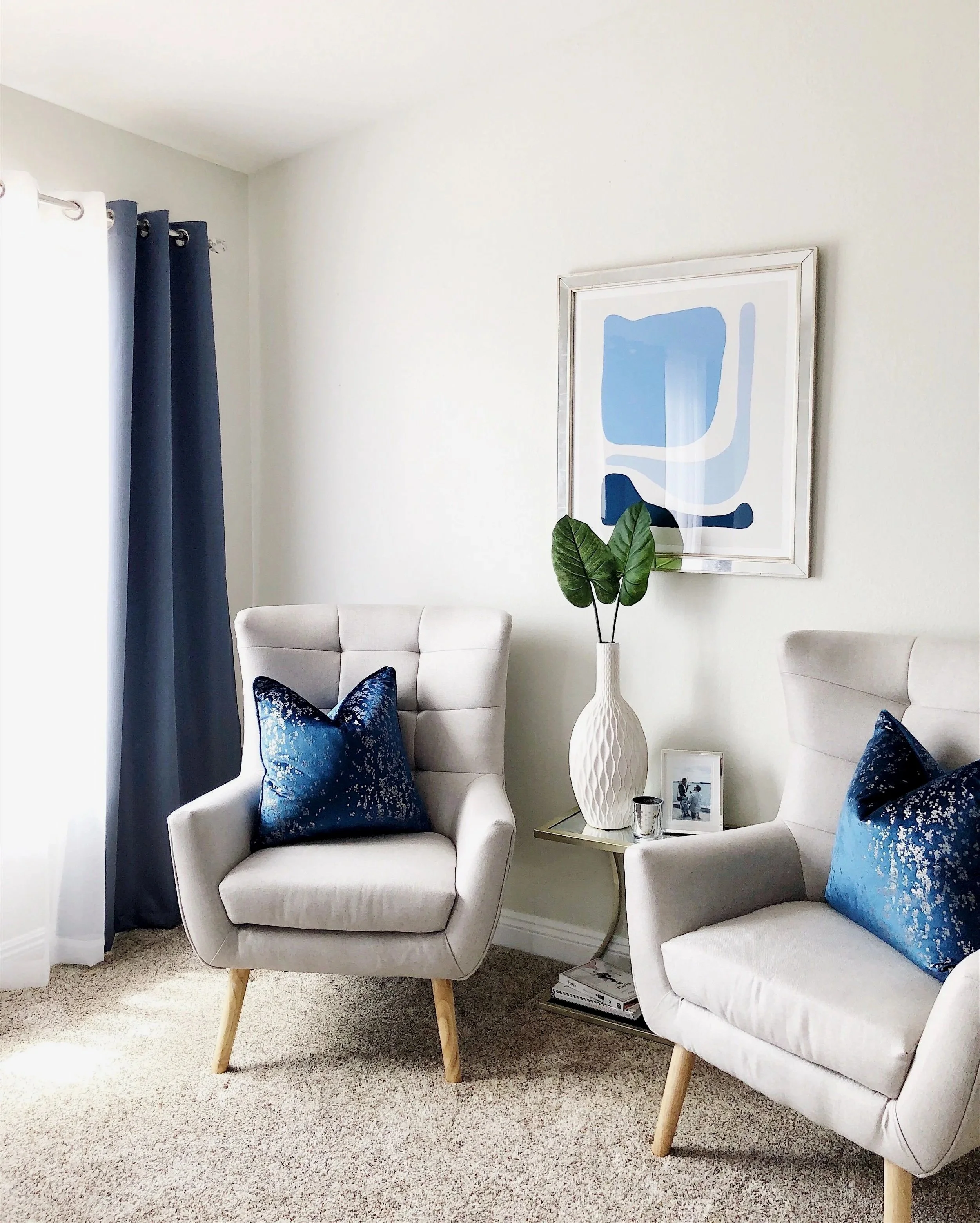 A cozy living room corner with two white armchairs with wooden legs, blue velvet throw pillows, a small side table with a white vase with green leaves, a framed abstract art piece on the wall, a framed photo on the side table, and dark blue curtains by the window.