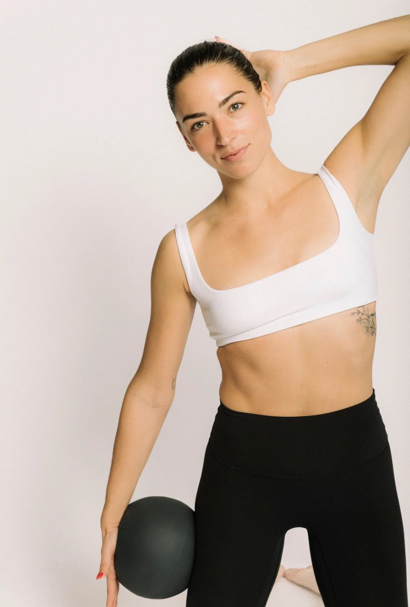 A woman with dark hair tied back, wearing a white sports bra and black leggings, holding a black exercise ball in her right hand and kneeling on the ground against a plain white wall.