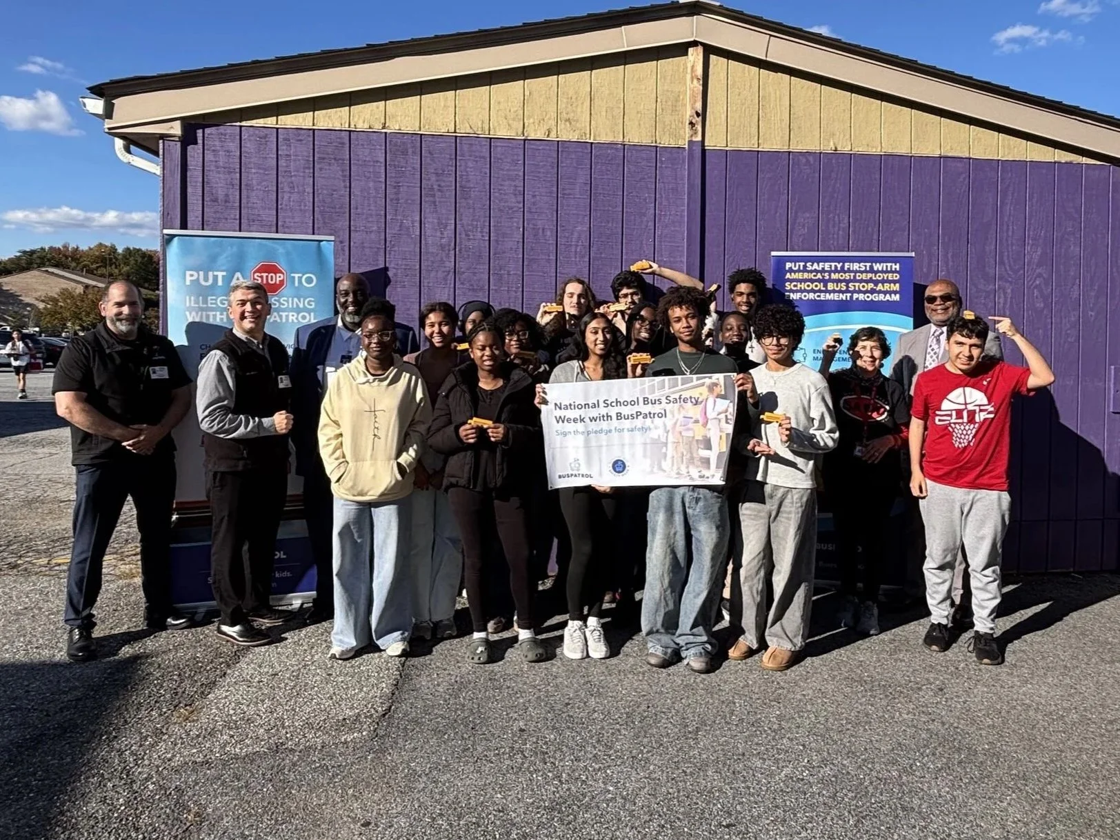 A group of people, including students and adults, gathered outside in front of a purple building, participating in National School Bus Safety Week with BusPatrol. They are holding a banner and some are holding small yellow school bus figures.