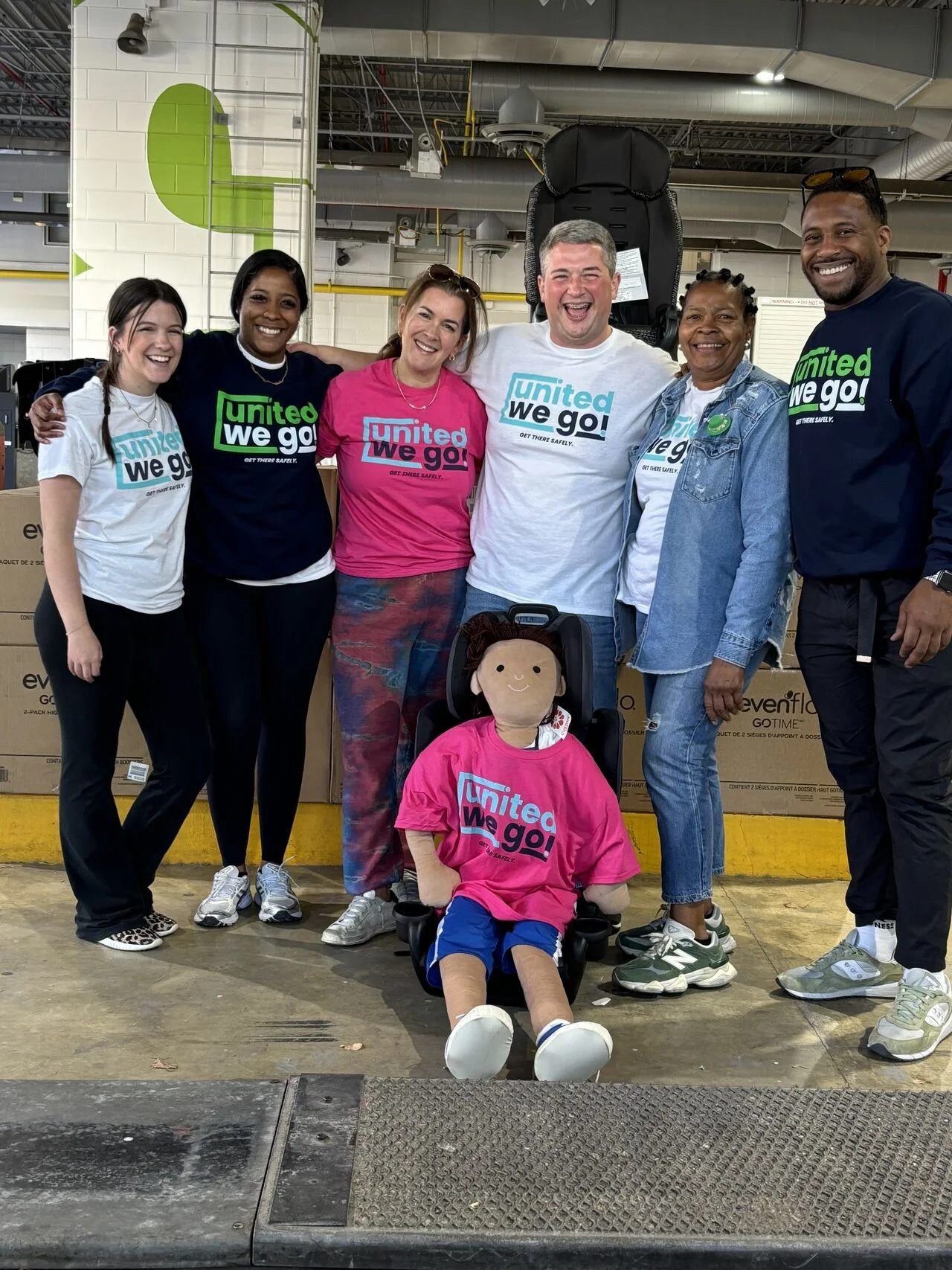 Group of six diverse people smiling and standing together at a child safety seat inspection station, all wearing shirts with the "United We Go!" slogan.