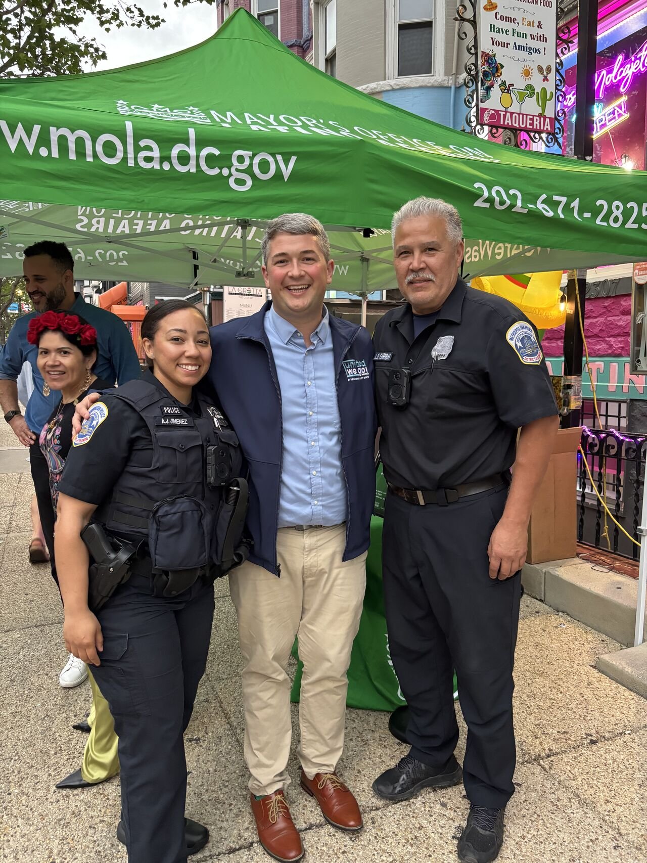 Three smiling people standing together outdoors, two police officers with badges and a man wearing a blue jacket, under a green tent with the website www.mola.dc.gov and a phone number, in front of a colorful storefront.