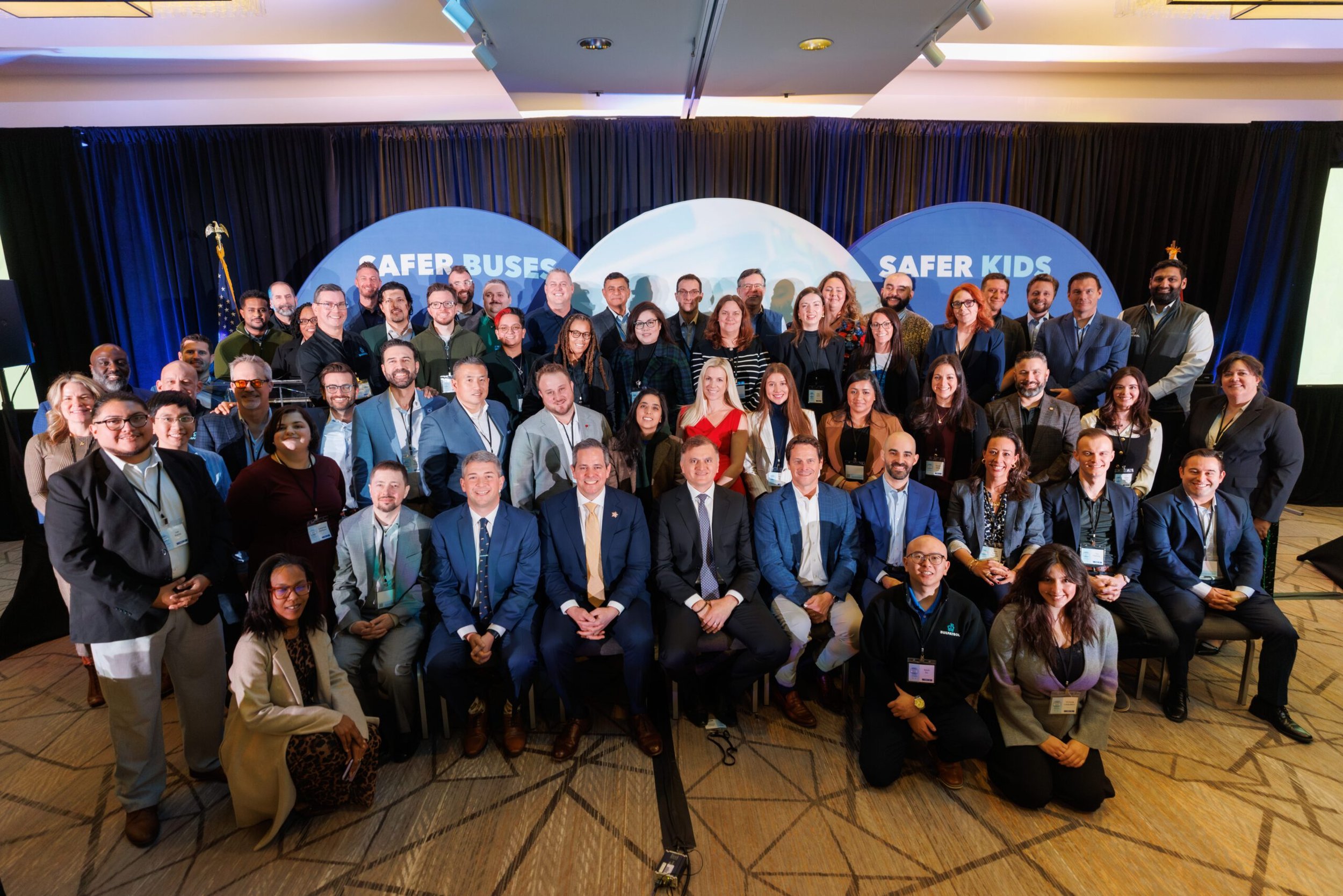 Group of diverse people at the 2026 National School Bus Safety Summit, standing in front of a stage with banners that say 'Safer Buses' and 'Safer Kids'.