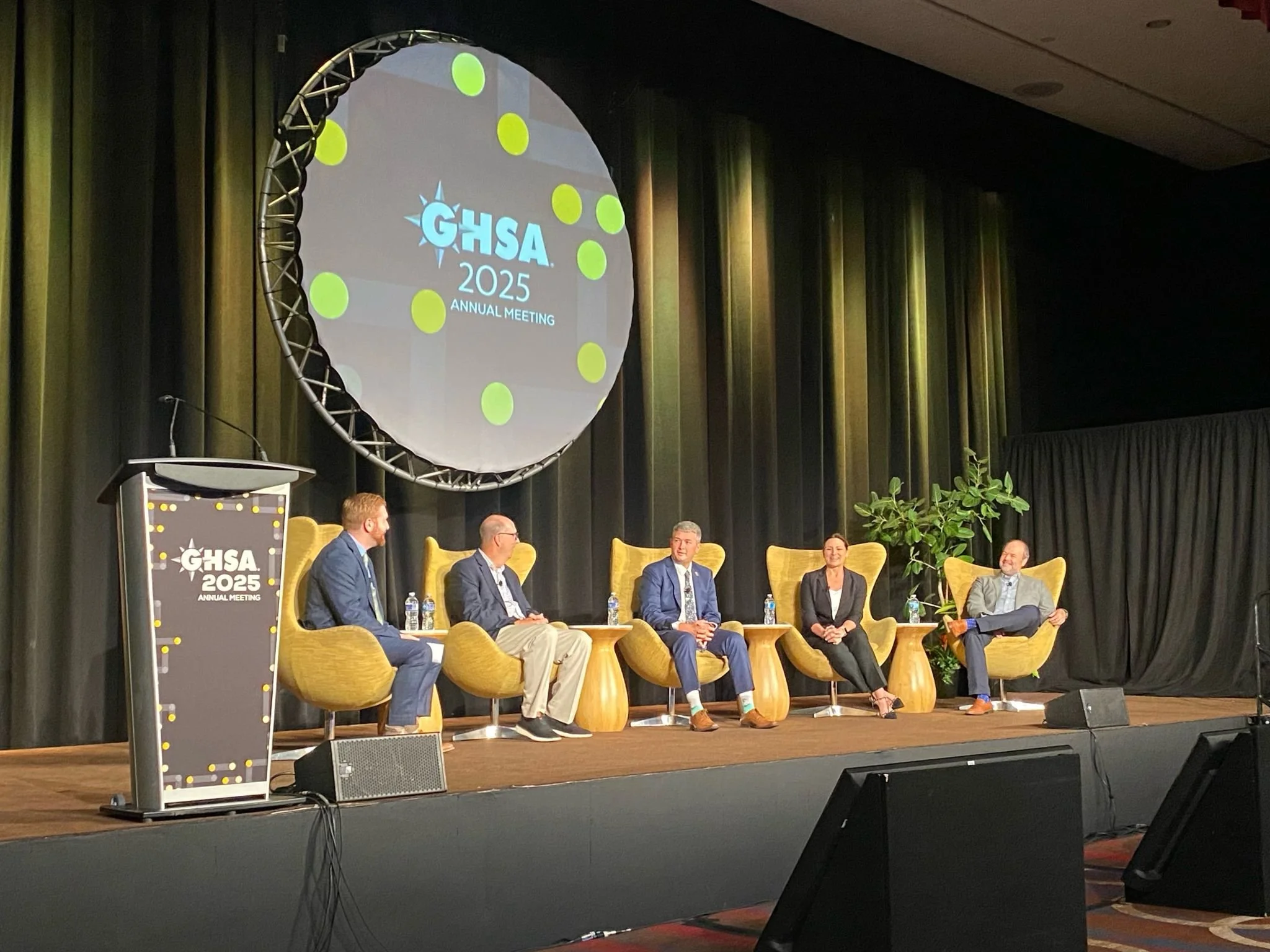 A panel of six individuals seated on stage at the GHSA 2025 Annual Meeting, with a large screen behind them displaying the event's logo and title, and a podium with the same logo in front of the stage.