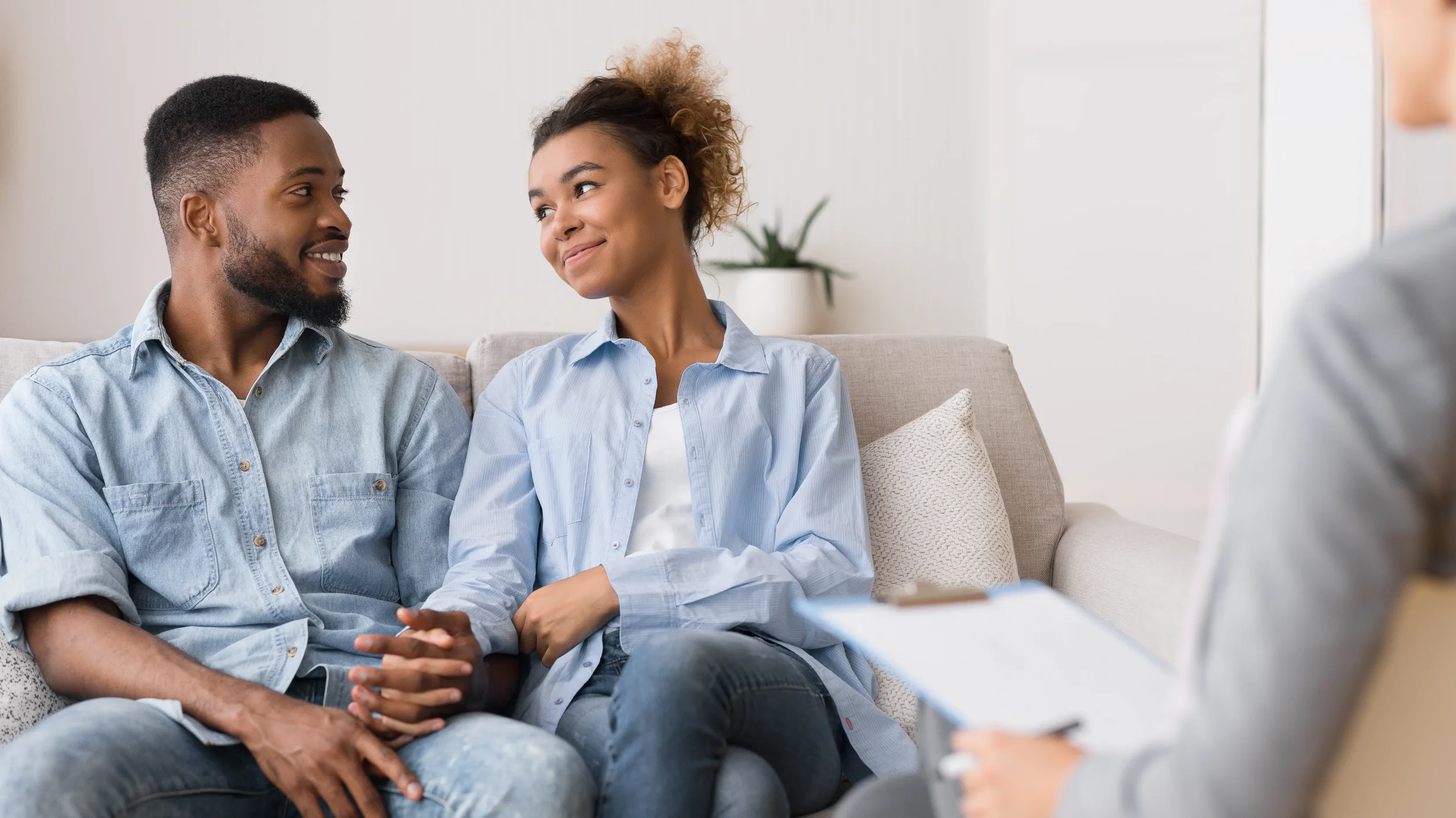 A man and woman sitting on a beige couch, smiling at each other during a conversation in a bright room with white walls and a potted plant in the background.
