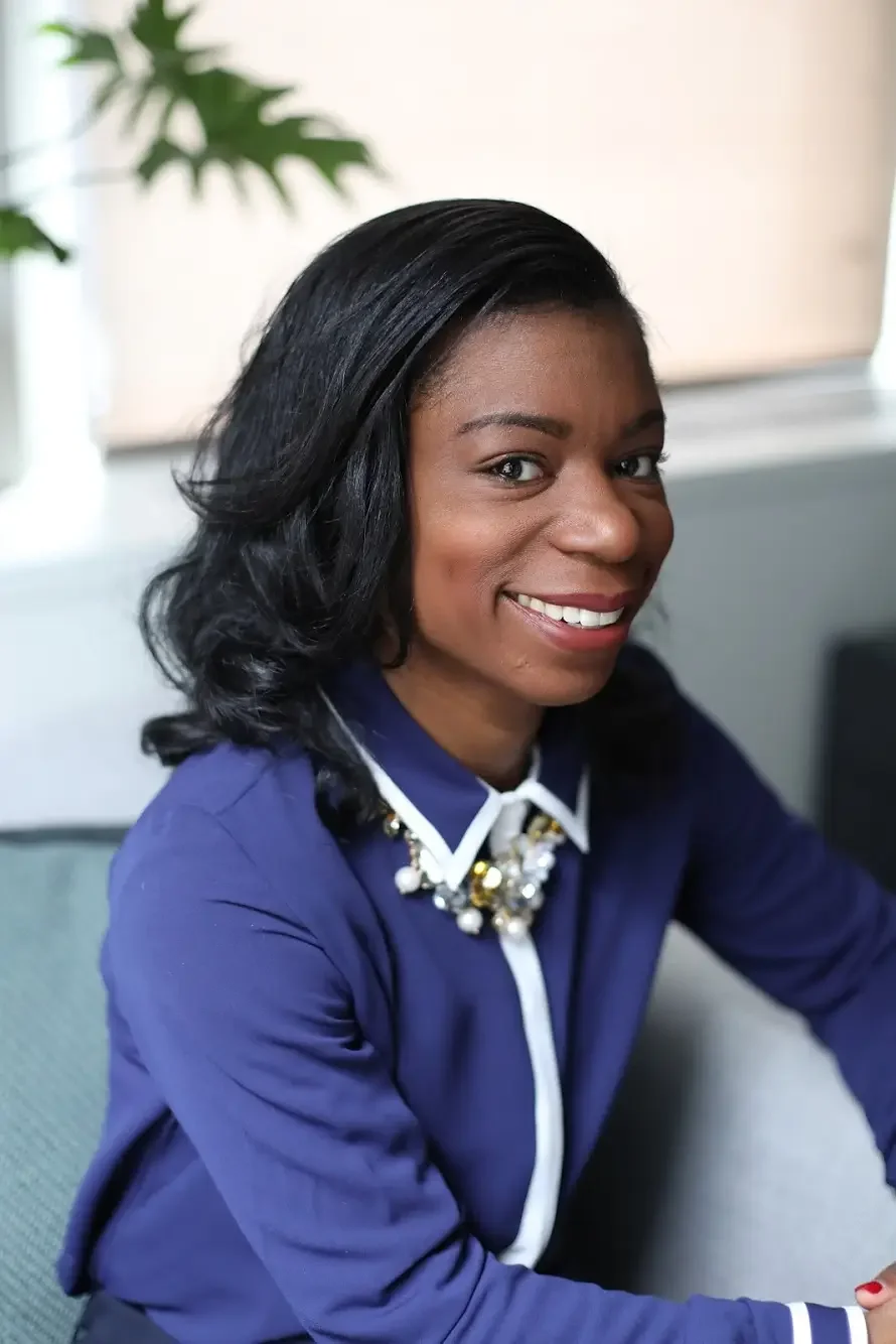 A smiling African American woman with black hair wearing a dark blue shirt and a decorative pearl necklace, sitting at a desk in an office environment.