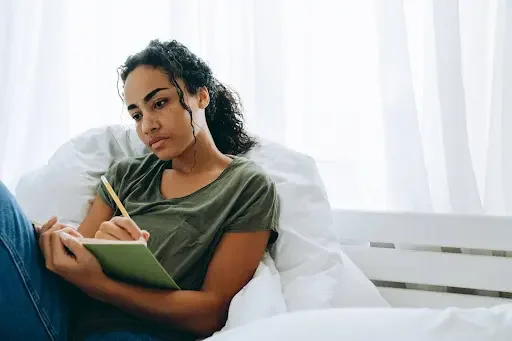 Young woman sitting on a bed, writing in a green notebook with a pencil, in a bright room with white curtains.