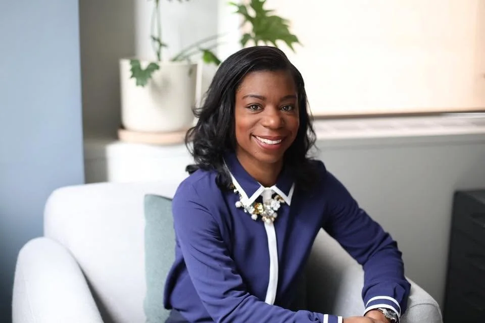 A smiling African American woman wearing a dark blue blouse, sitting on a light-colored chair in a well-lit room
