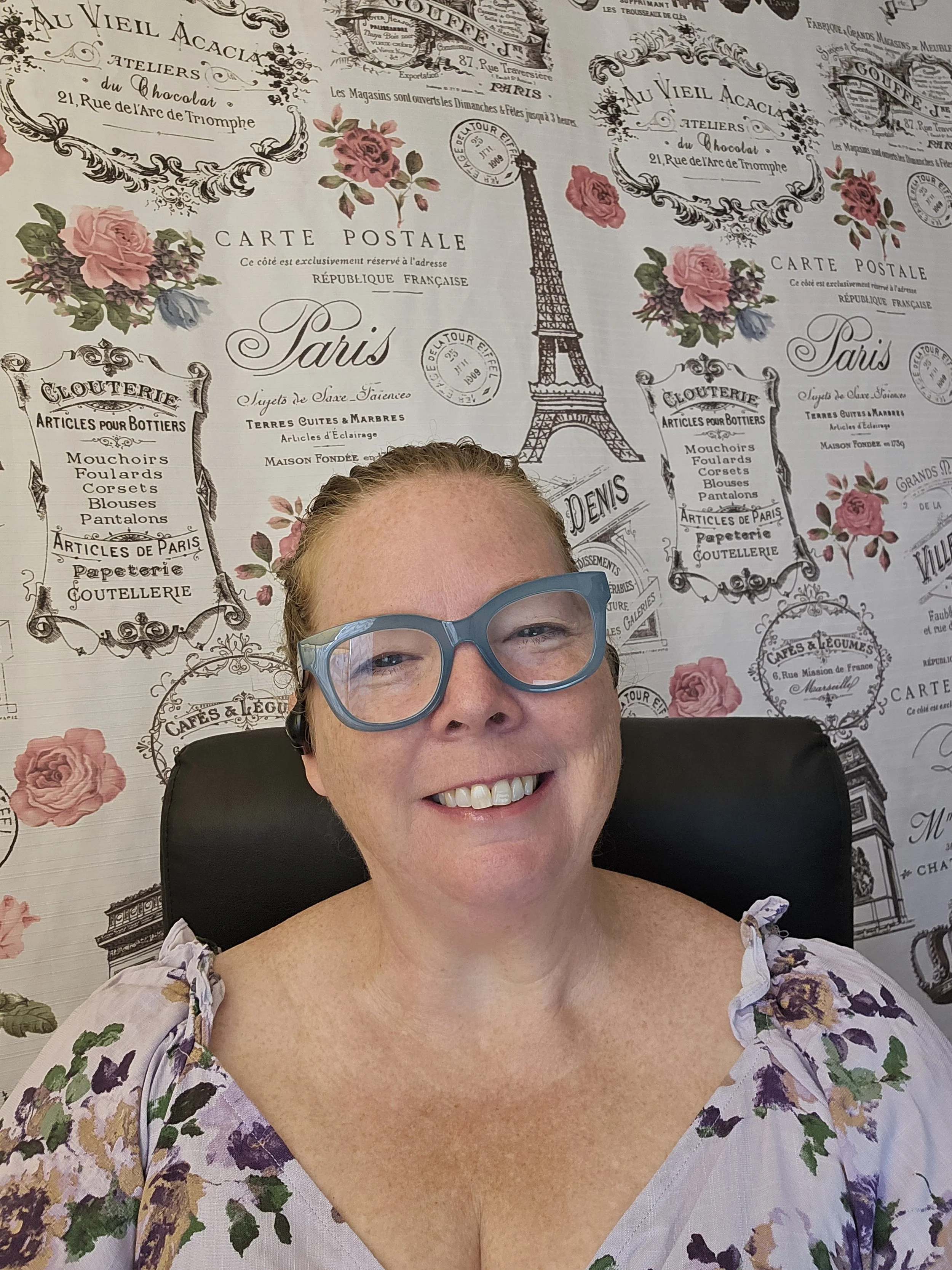 Close-up of a smiling woman wearing large blue glasses, seated in front of a decorative Paris-themed wallpaper with images of the Eiffel Tower and floral designs.