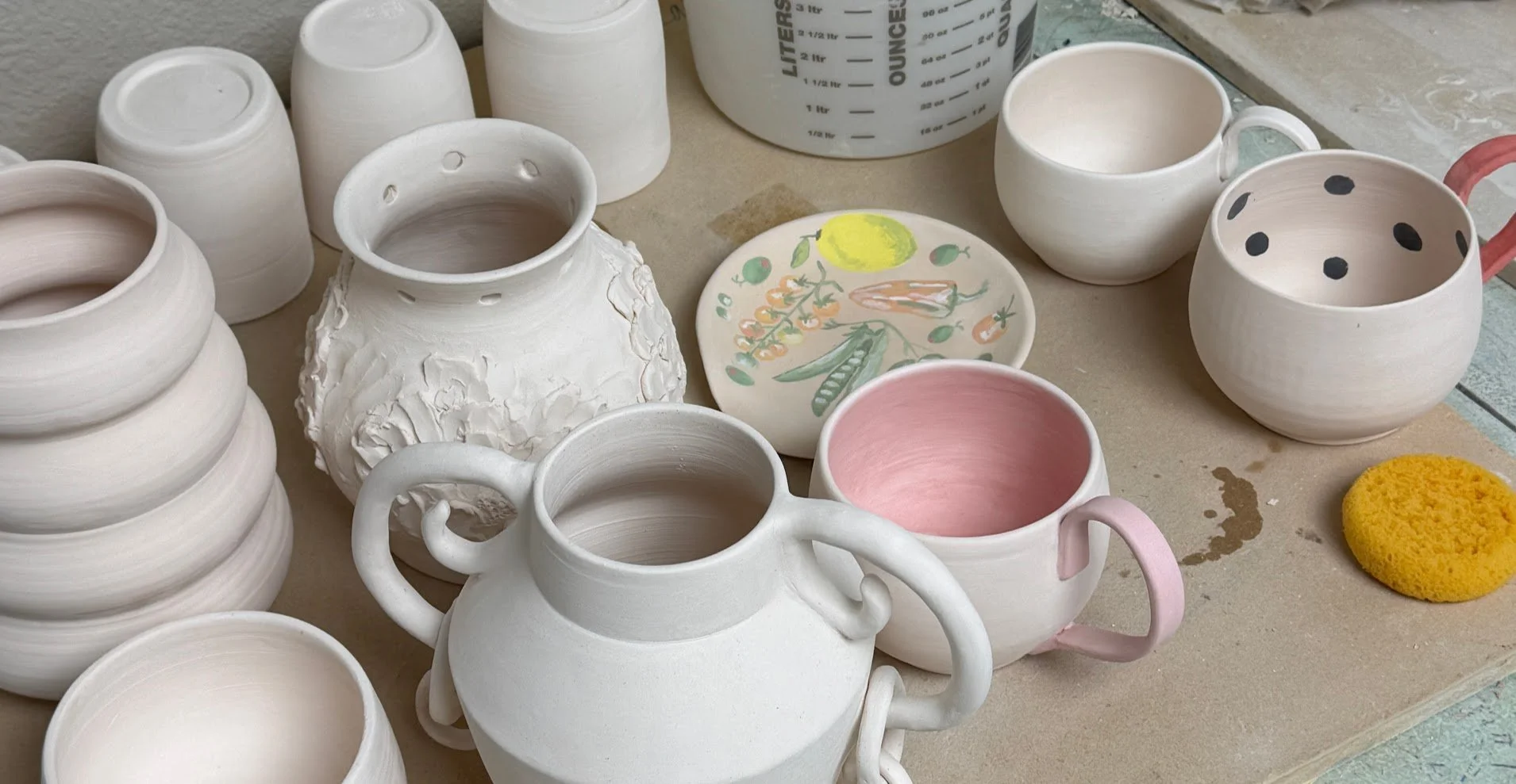 Unfinished white ceramic pottery items, including cups, a teapot, and bowls, with some featuring painted designs, on a worktable with a sponge and a painted plate.