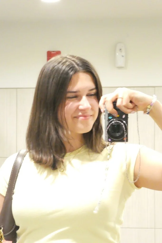 A young woman taking a mirror selfie in a room with beige walls, wearing a light yellow shirt and a bracelet on her right wrist.