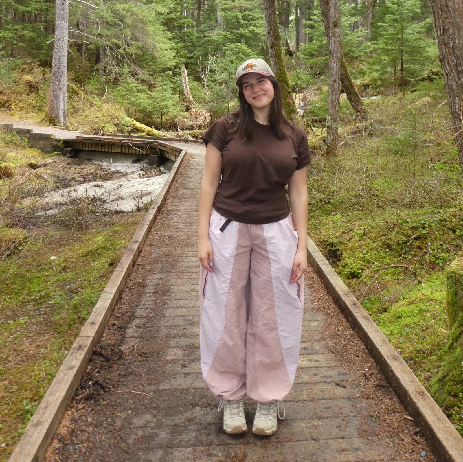 A young woman standing on a wooden trail in a forest during daytime, smiling and wearing a cap, brown T-shirt, and pink pants with white sneakers.