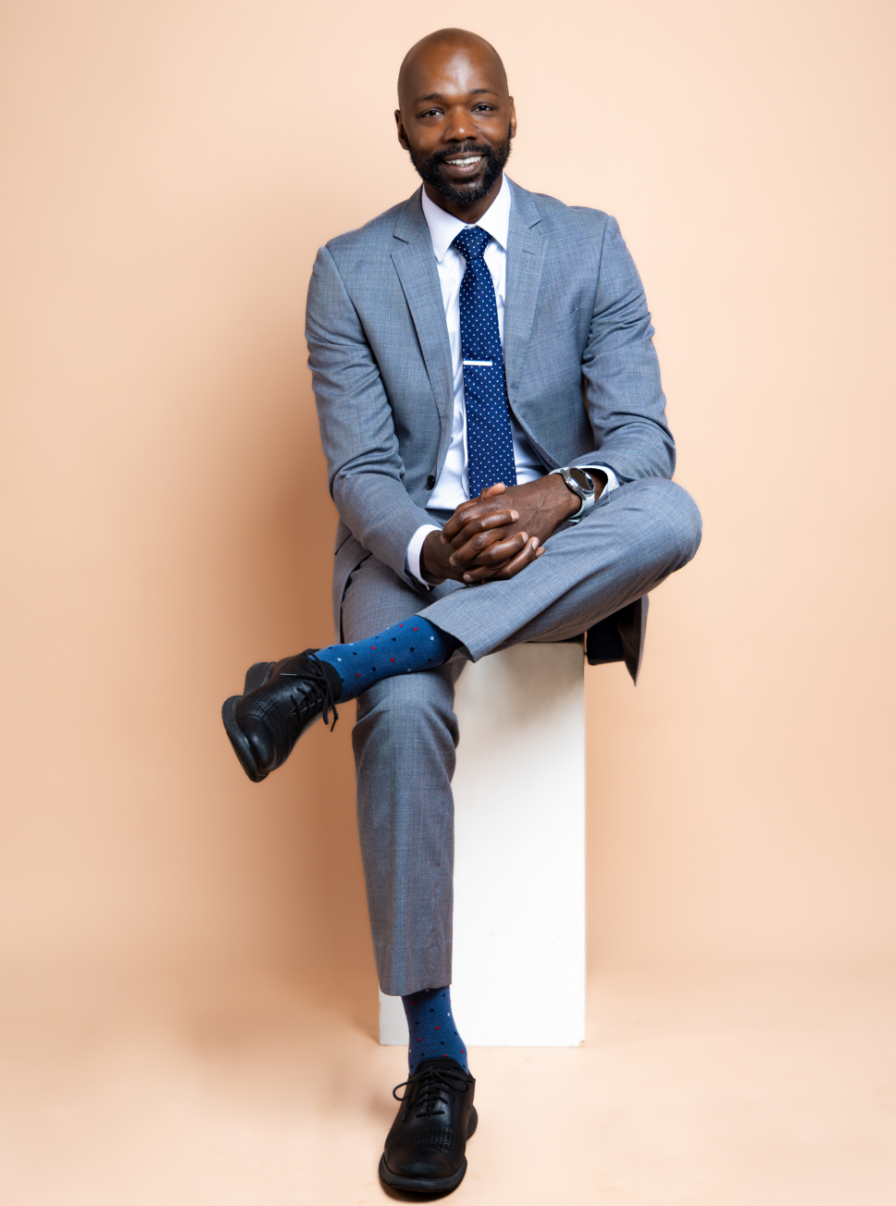 A man in a gray suit, white shirt, and blue tie sitting on a white stool against a beige background. He has a beard, is smiling, and has his hands clasped on his knee, with one leg crossed over the other. He is wearing black shoes and blue socks with red dots.