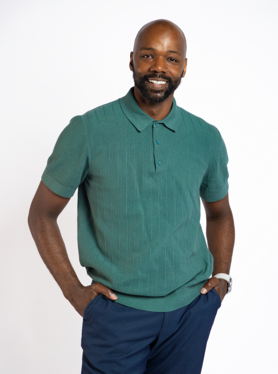 A smiling man with a shaved head and beard wearing a green polo shirt and navy pants, posing with one hand in his pocket against a white background.