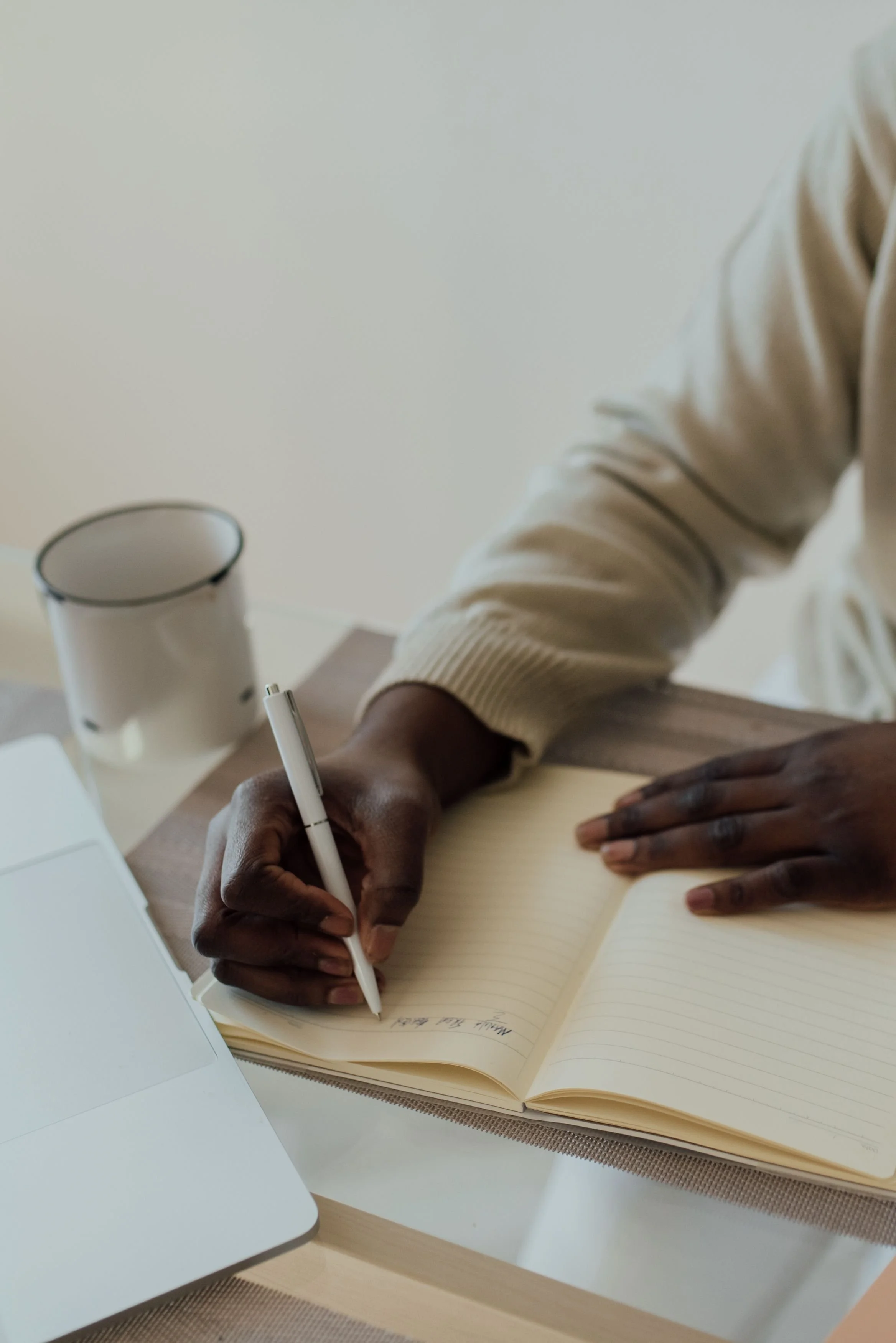 Two people with dark skin are working together at a desk. One person is writing in a notebook with a white pen, and the other person is resting their hand on the open notebook. There is a white mug, a laptop, and a tablet on the desk.