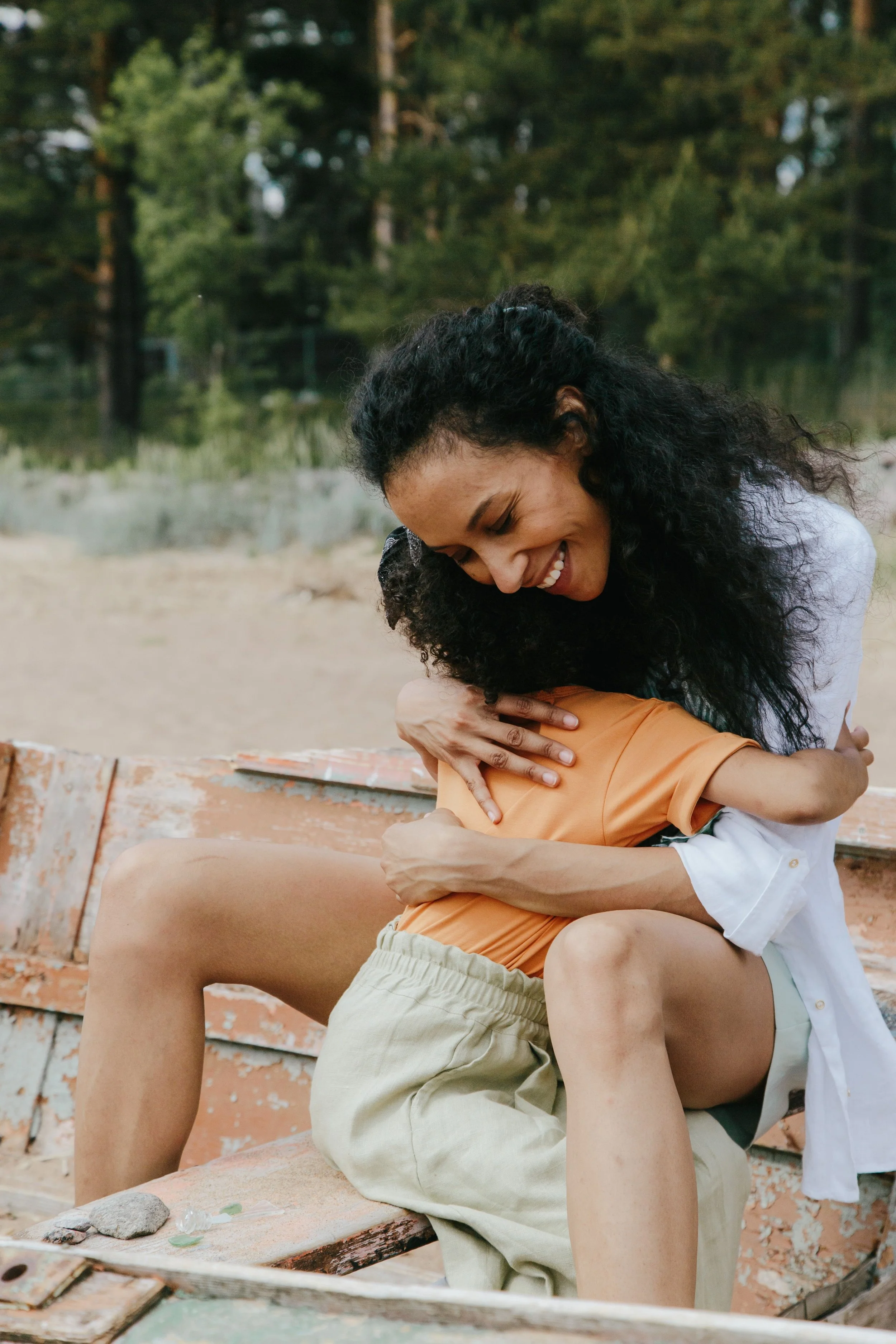 A young woman with curly hair smiling and hugging a person in shorts outdoors, sitting on a weathered wooden bench with a background of trees and a grassy field.