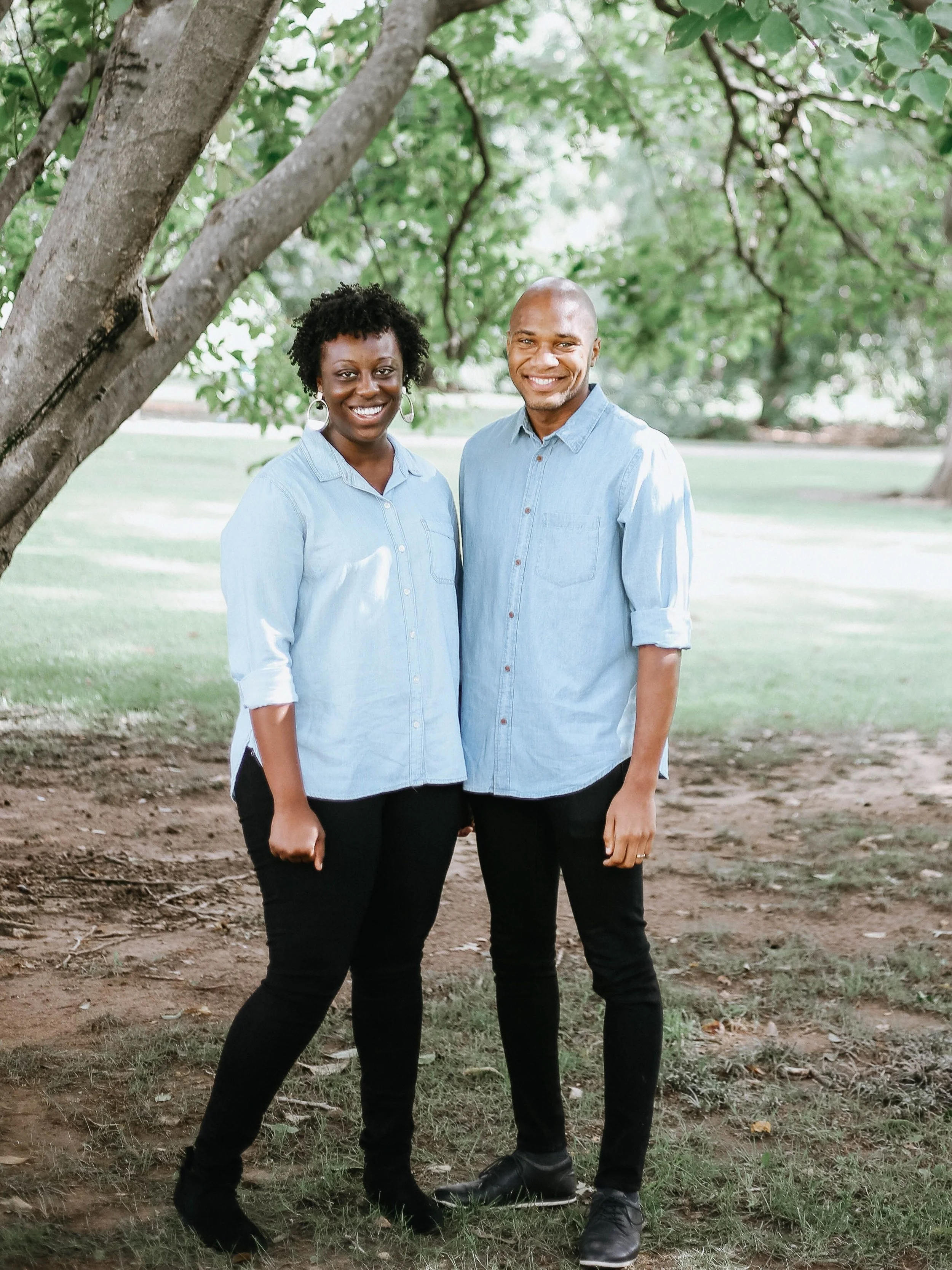 A smiling African American woman and a smiling African American man are standing together outdoors under a tree in a park. Both are wearing light blue denim shirts and dark pants.