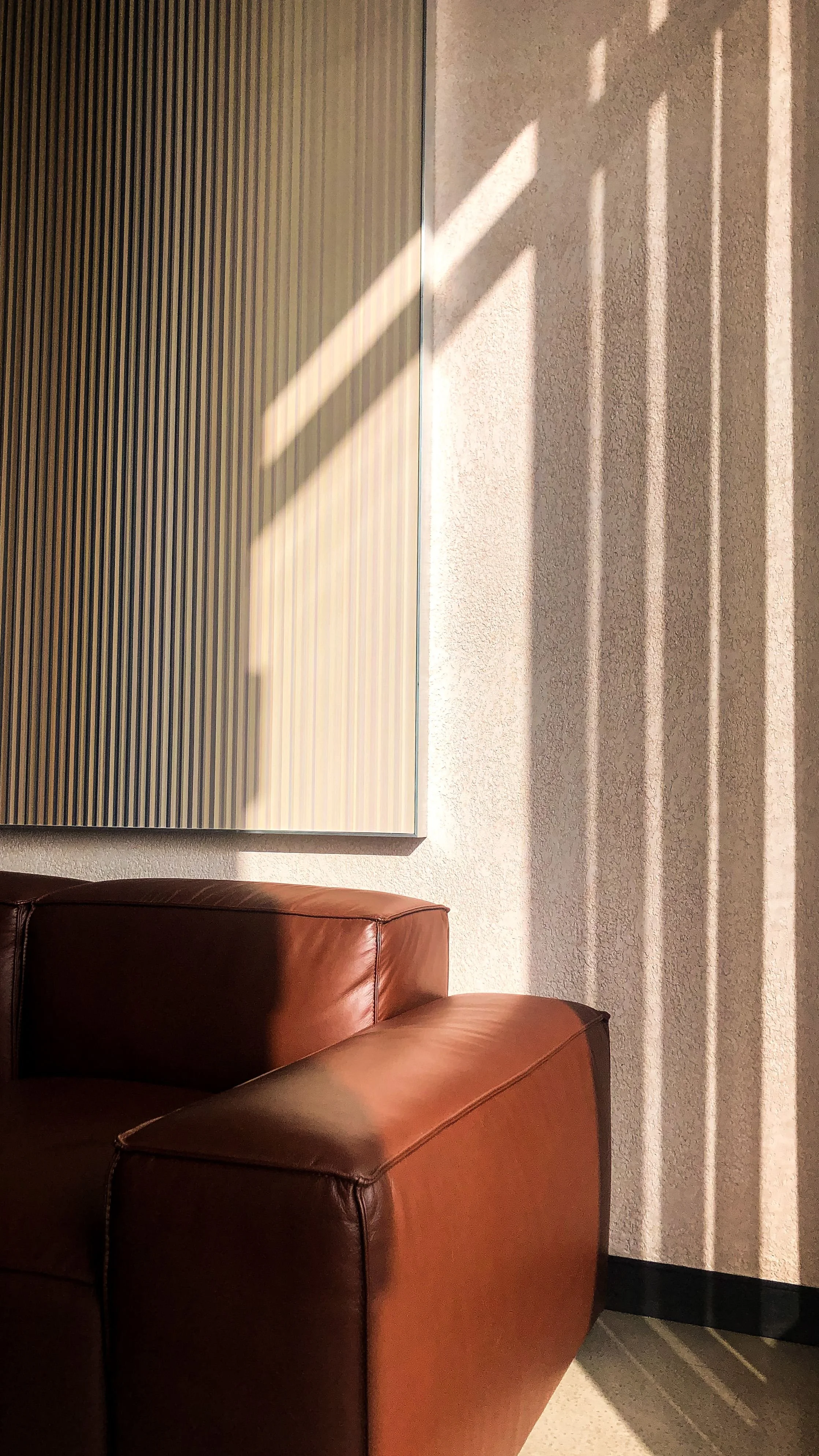 Sunlight casts shadows through window blinds onto a wall and part of a leather armchair in a room.