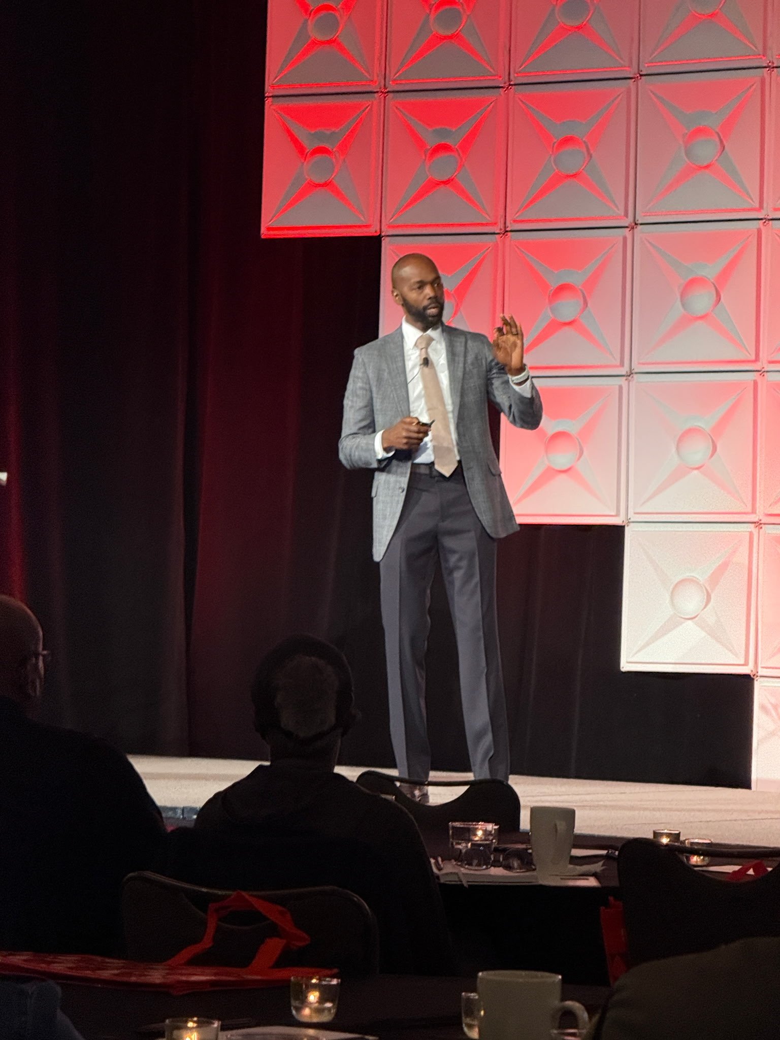 A man in a gray suit and beige tie giving a presentation on a stage with a red geometric backdrop, audience members seated at tables in front.
