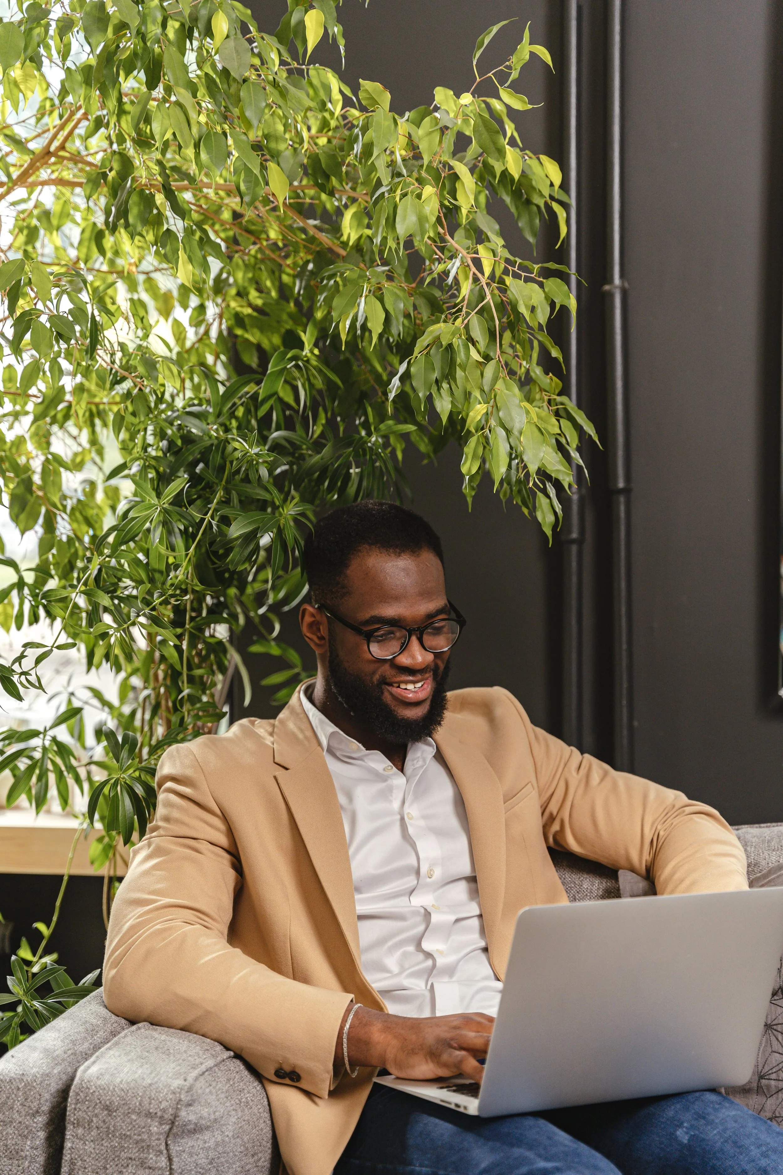 A man with glasses and a beard, wearing a beige blazer and white shirt, sitting on a gray couch with a laptop. There are green plants behind him and a dark wall with black pipes.