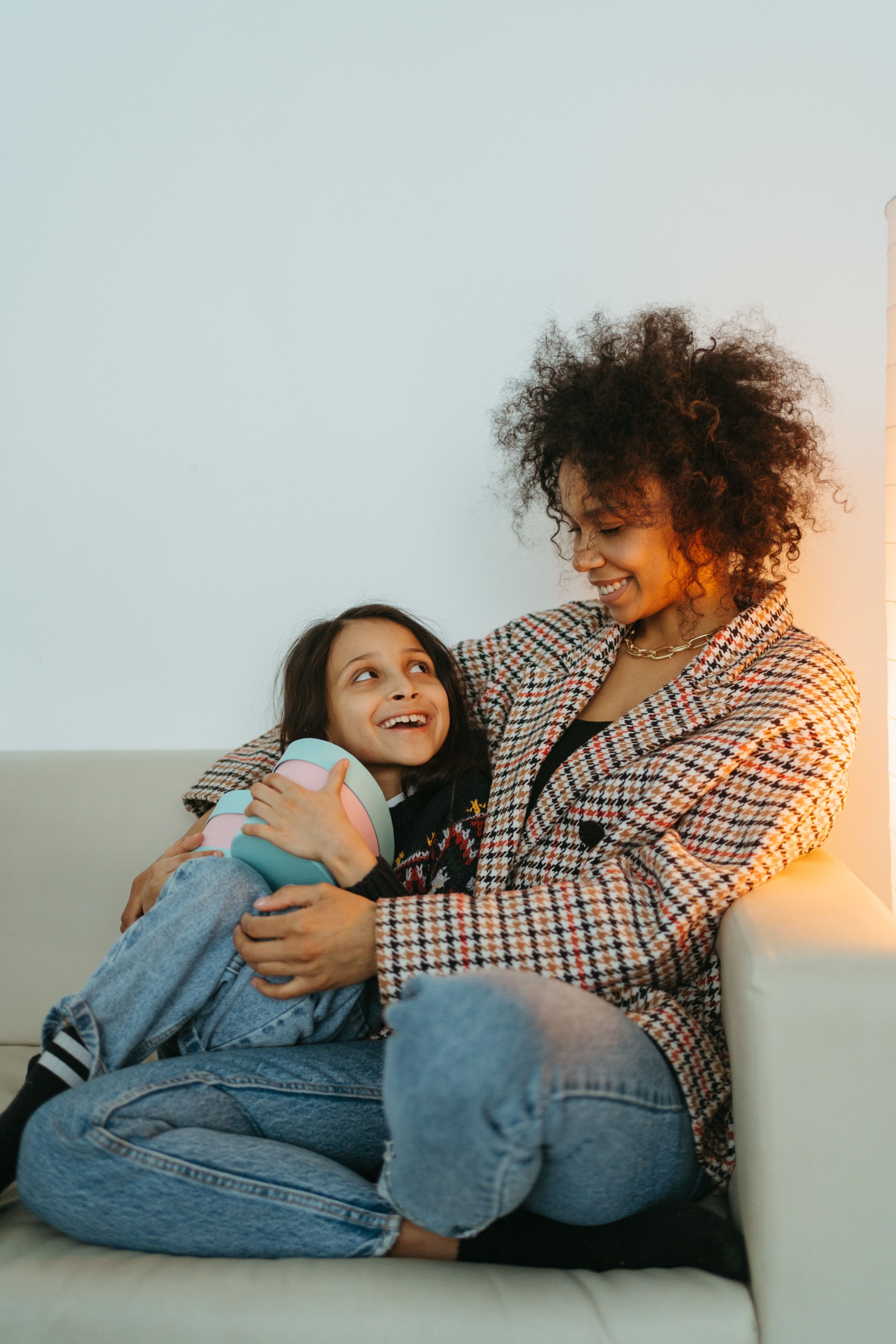 A woman and a girl sitting on a white couch, smiling and embracing each other, with the woman looking affectionately at the girl in a cozy indoor setting.