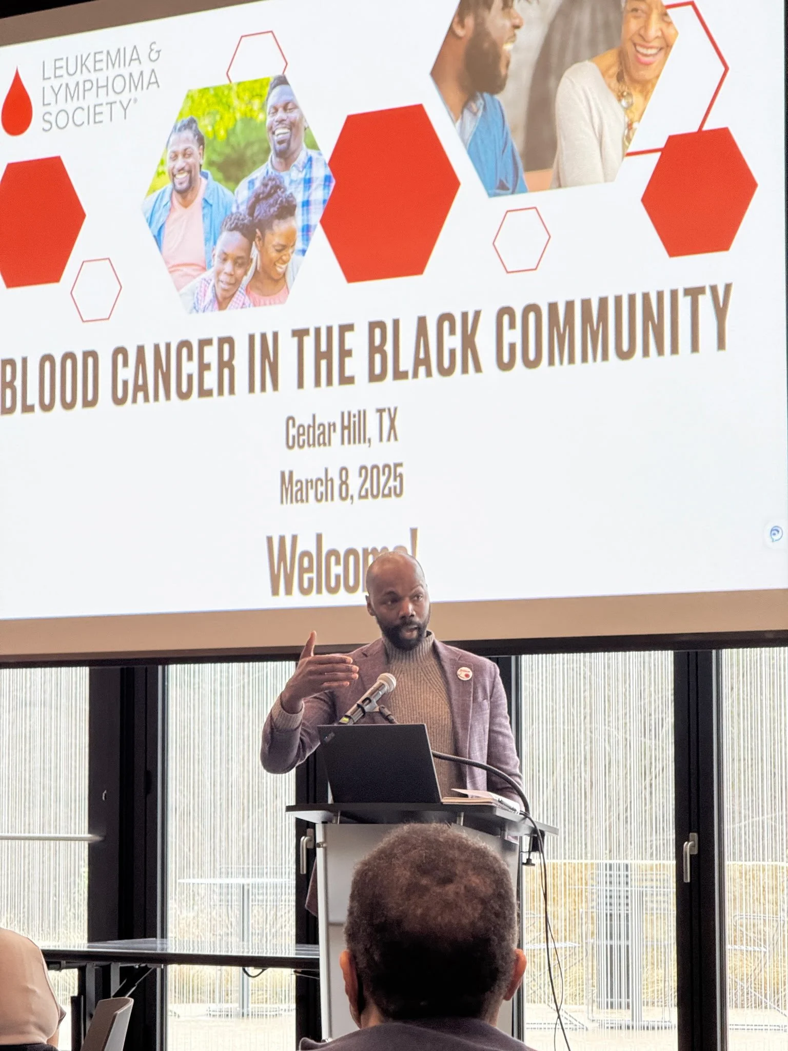 A man with a beard and mustache, wearing a gray blazer and beige turtleneck sweater, is giving a presentation at a conference about blood cancer in the Black community. He is standing behind a podium with a microphone, gesturing with his right hand, and looking at the audience. In the background, there is a large screen displaying a slide with the title 'Blood Cancer in the Black Community,' the location 'Cedar Hill, TX,' and the date 'March 8, 2025.' The slide also features the logos of the Leukemia & Lymphoma Society, the slide has images of smiling Black people.