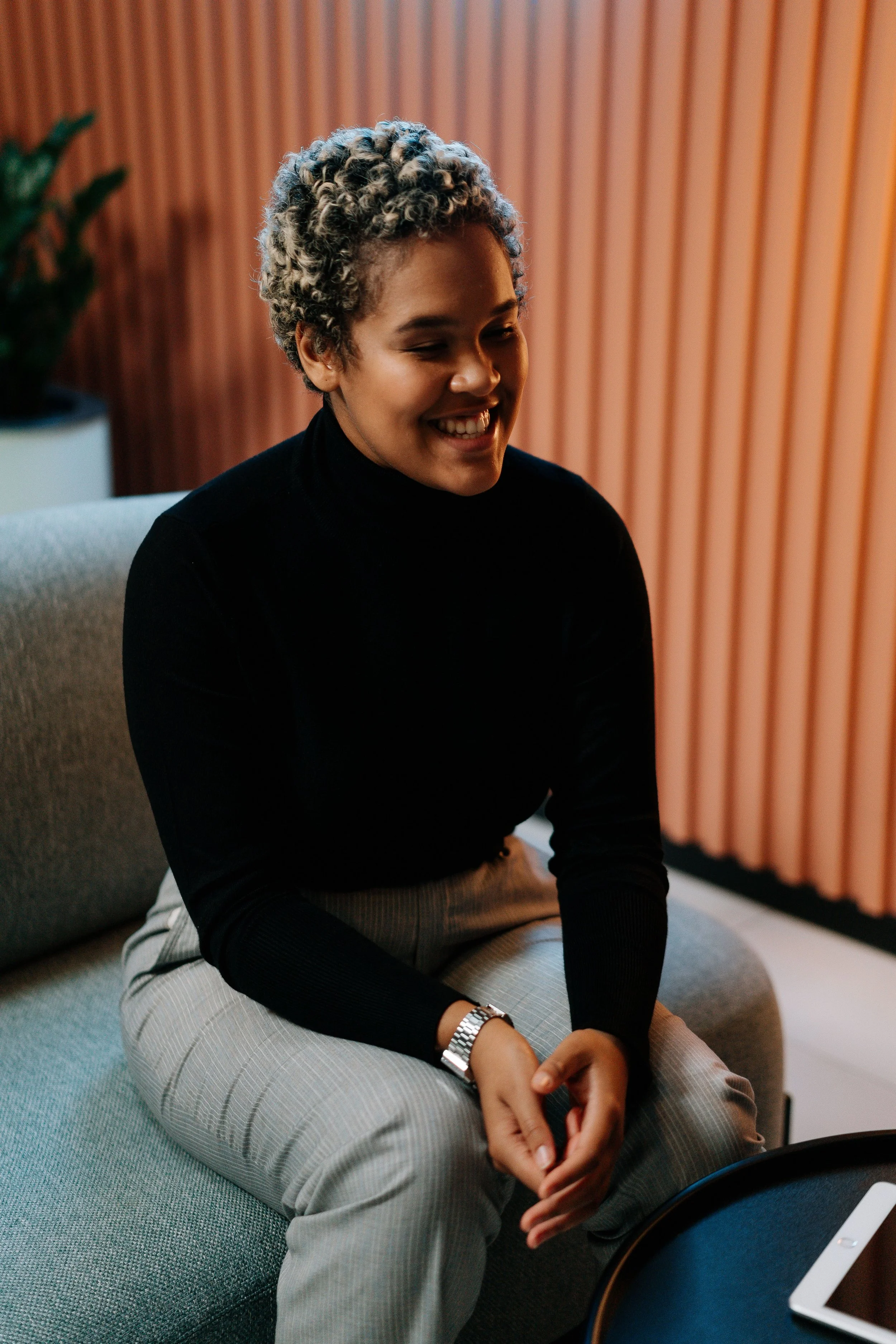 A woman sitting on a gray cushioned bench, smiling and looking down, wearing a black turtleneck, beige striped pants, and a silver watch, with a wooden wall and a potted plant in the background.