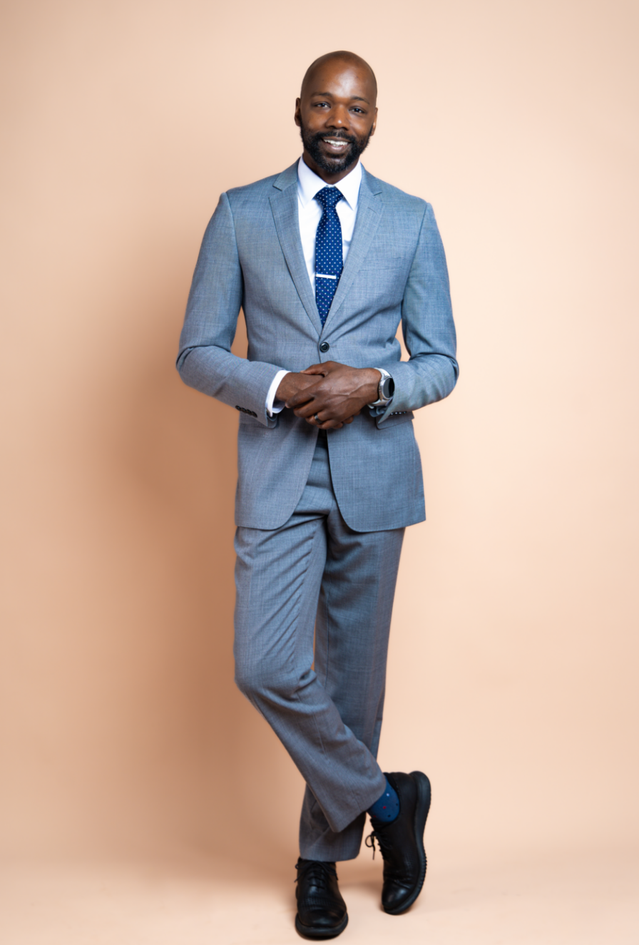 A well-dressed man in a light grey suit with a blue tie, white shirt, black shoes, and a watch, standing against a beige background, smiling with conference badge clipped to his pocket.