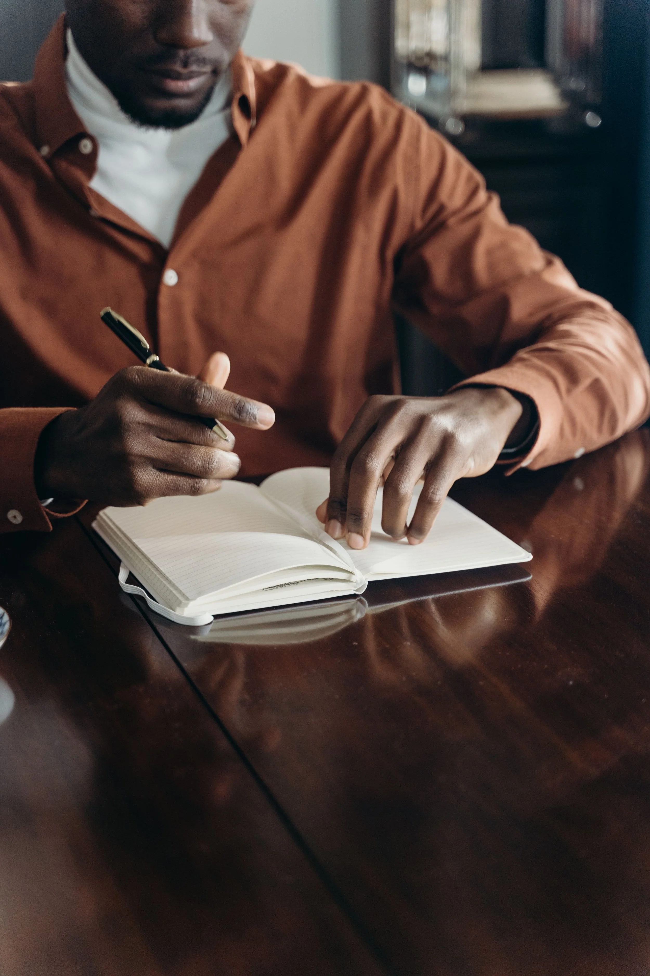 A man wearing a brown shirt and a white turtleneck is sitting at a dark wood table, writing in a lined notebook with a black pen. There is a blurred bookshelf in the background.