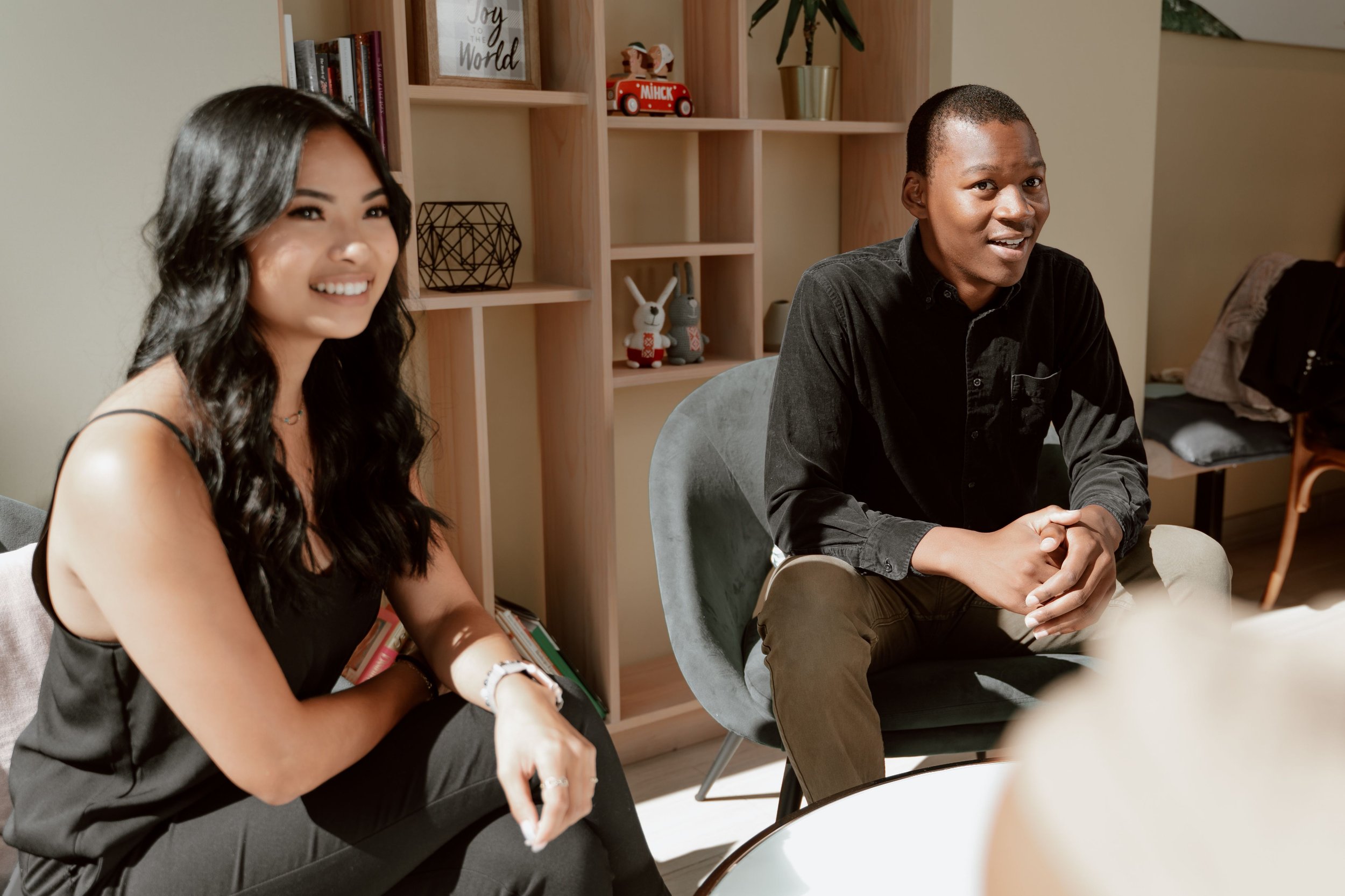 Two people sitting at a table, smiling. The woman on the left has long dark hair, wearing a black sleeveless top, and the man on the right has short hair, wearing a black shirt. Behind them is a wooden shelf with books and decorative items.