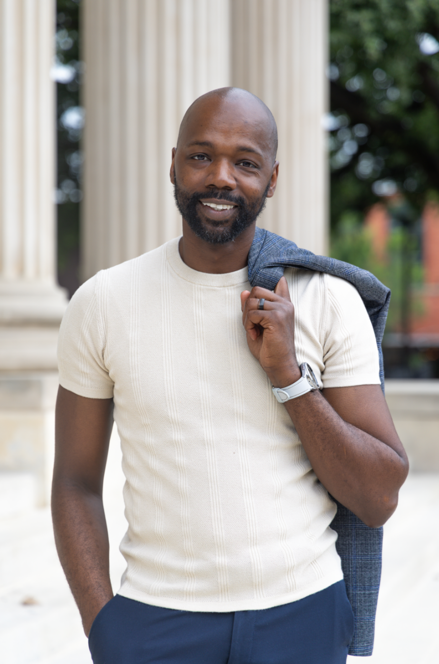 A smiling man with a beard and bald head carrying a jacket over his shoulder, standing outdoors near classical columns.