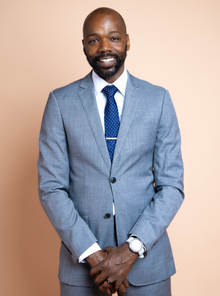 A man wearing a light gray suit, white shirt, and navy polka dot tie posing in front of a beige background.