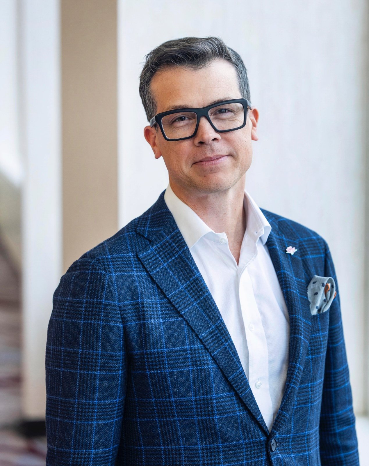 A man wearing glasses, a white shirt, and a blue plaid blazer with a pocket square, standing indoors near a light-colored wall.
