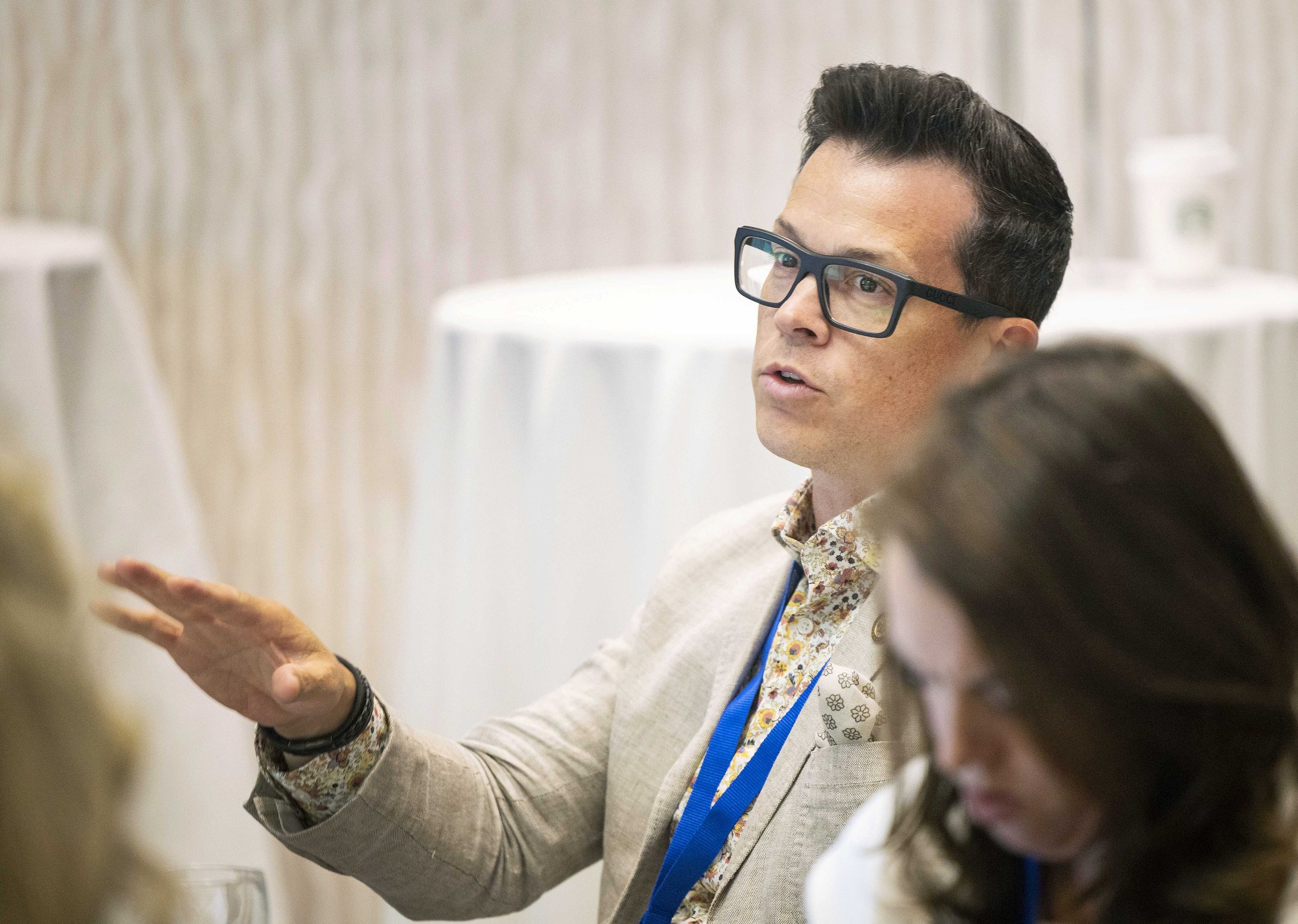 A man with dark hair, glasses, and wearing a beige blazer with a floral shirt, is speaking in a meeting or conference room.