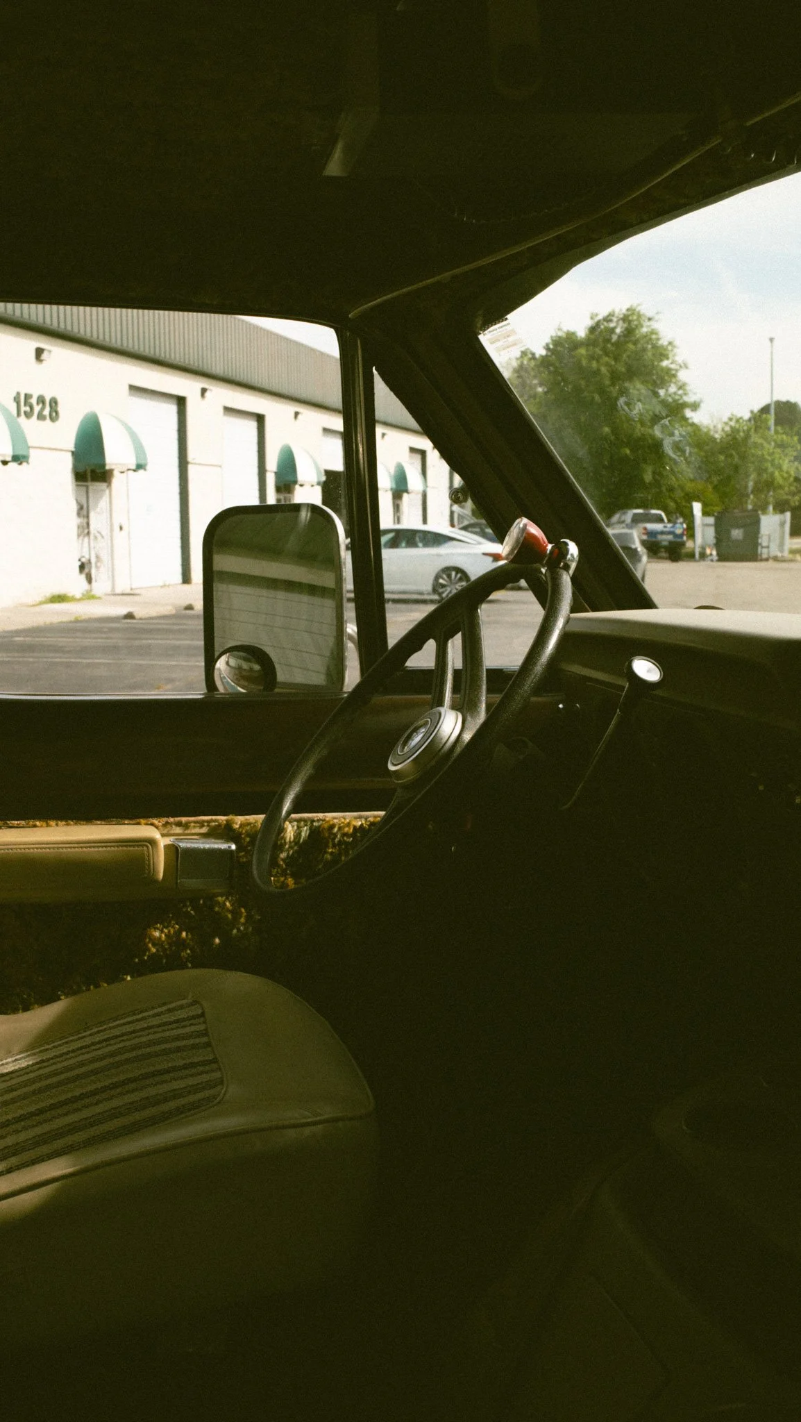 View from inside a vehicle showing the dashboard and steering wheel with the side mirror visible. Outside, a parking lot with a building, cars, trees, and a trash can can be seen.