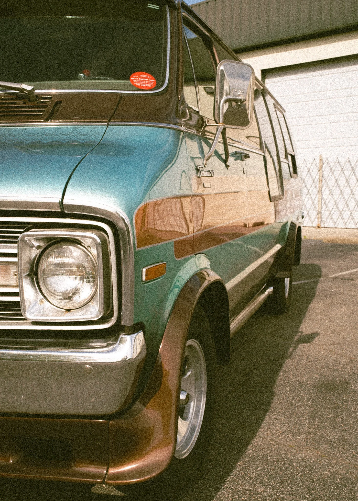 Close-up of the front right side of a vintage multicolored van with a black roof and turquoise, orange, and brown body, parked on a paved area near a building with a white garage door and chain-link fence.