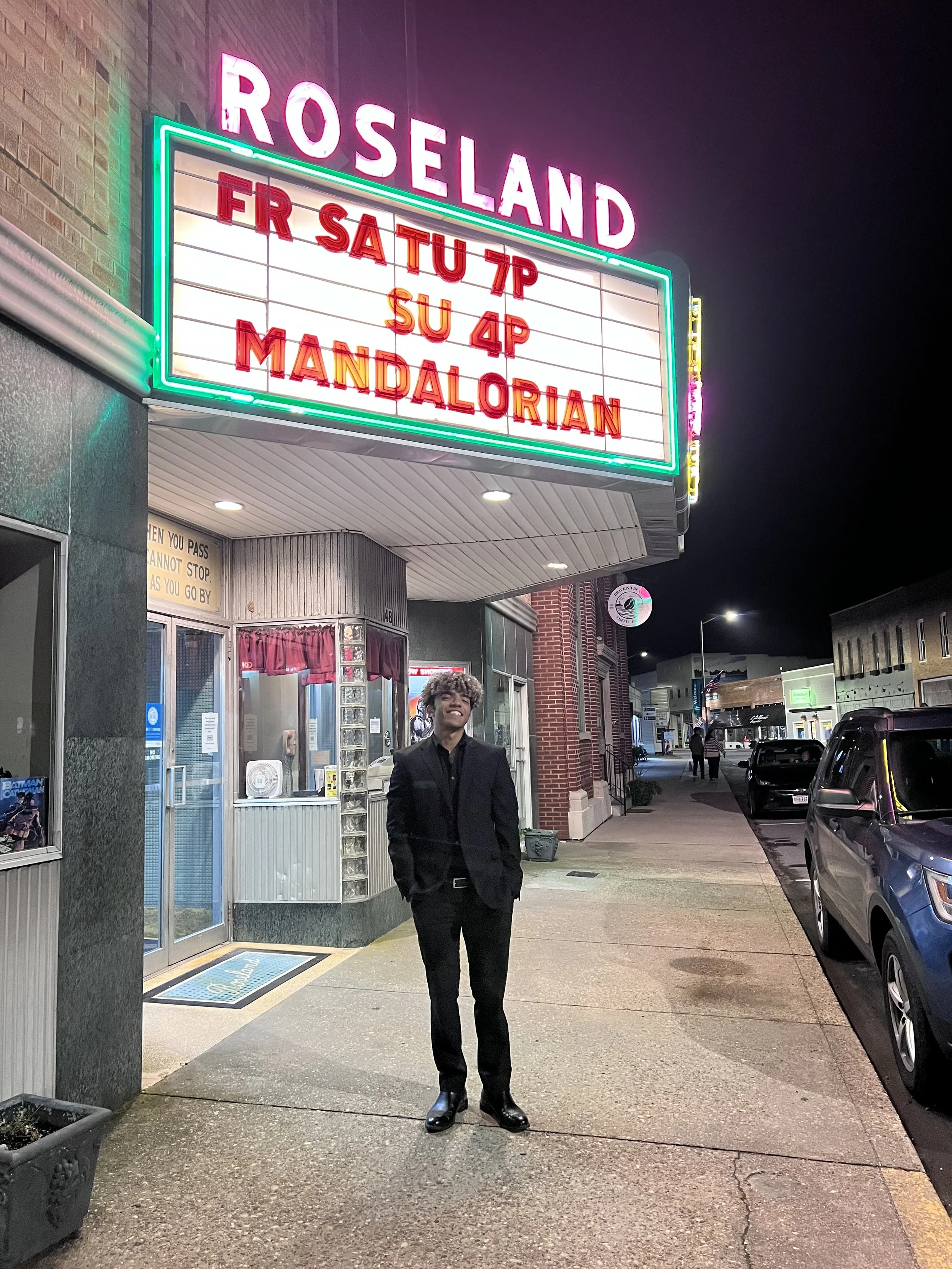 A man in a black suit standing outside a neon-lit movie theater at night. The theater sign says 'ROSELAND' at the top, with movie titles listed below. The street has parked cars and some pedestrians in the background.