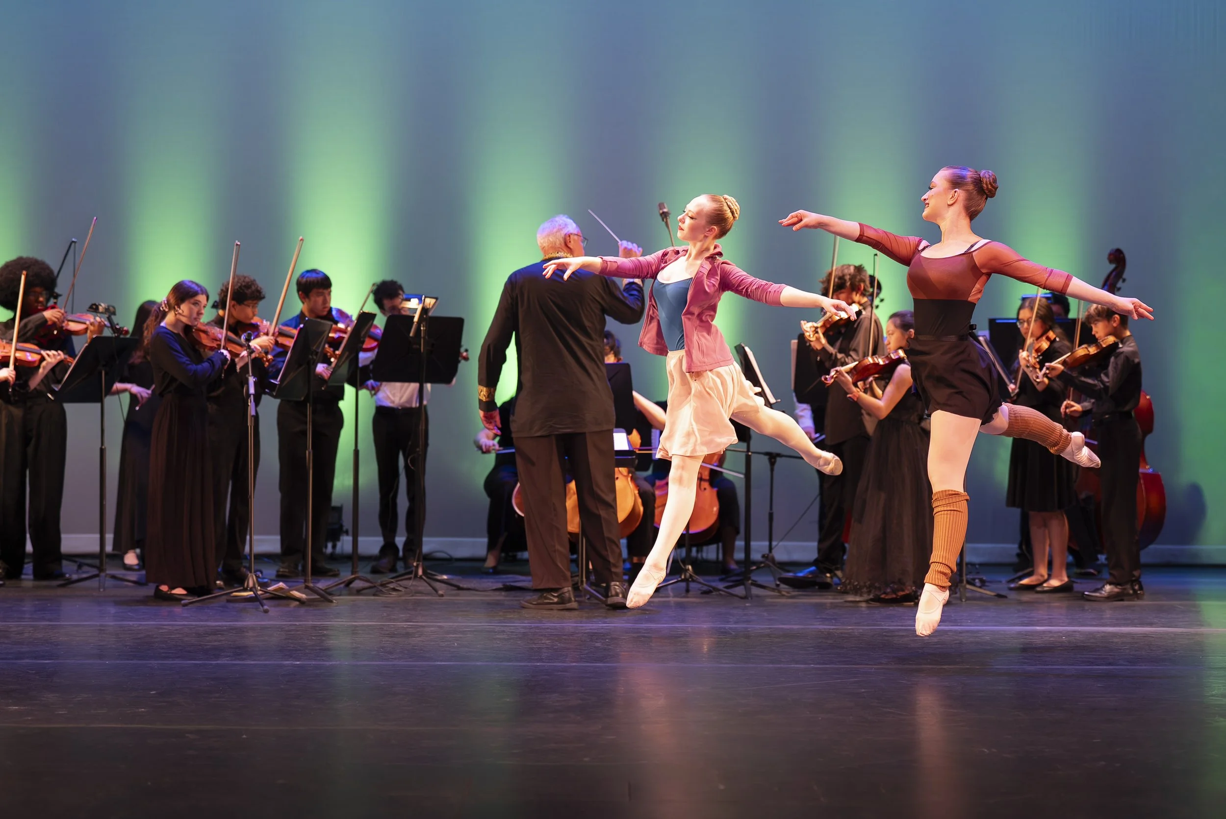 Ballet dancers performing on stage in front of an orchestra with green lighting background.