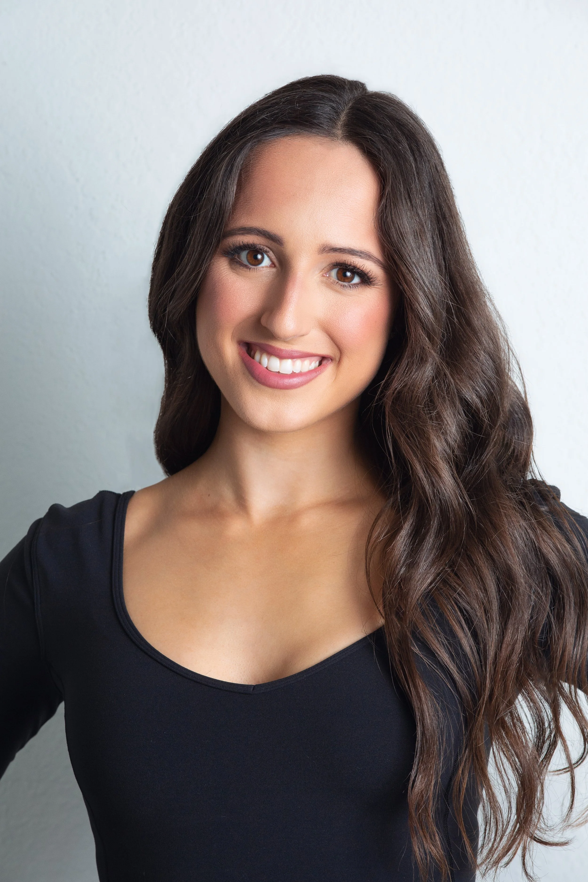 Close-up portrait of a young woman with long, wavy brown hair, brown eyes, and a bright smile, wearing a black top, against a plain white background.