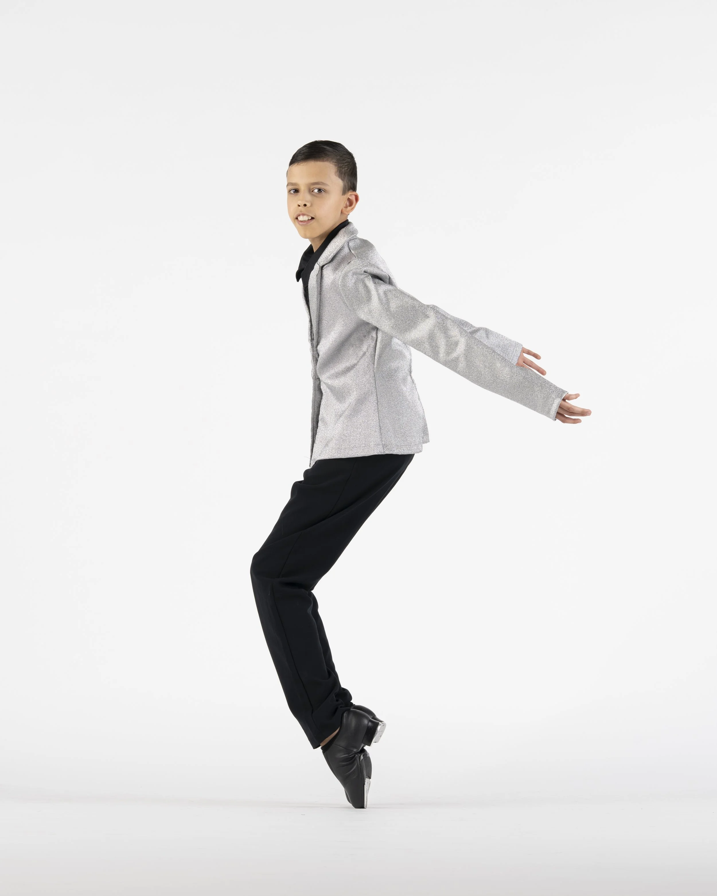 A boy in a silver jacket and black pants figure skating on ice, with arms extended and balancing on one skate.