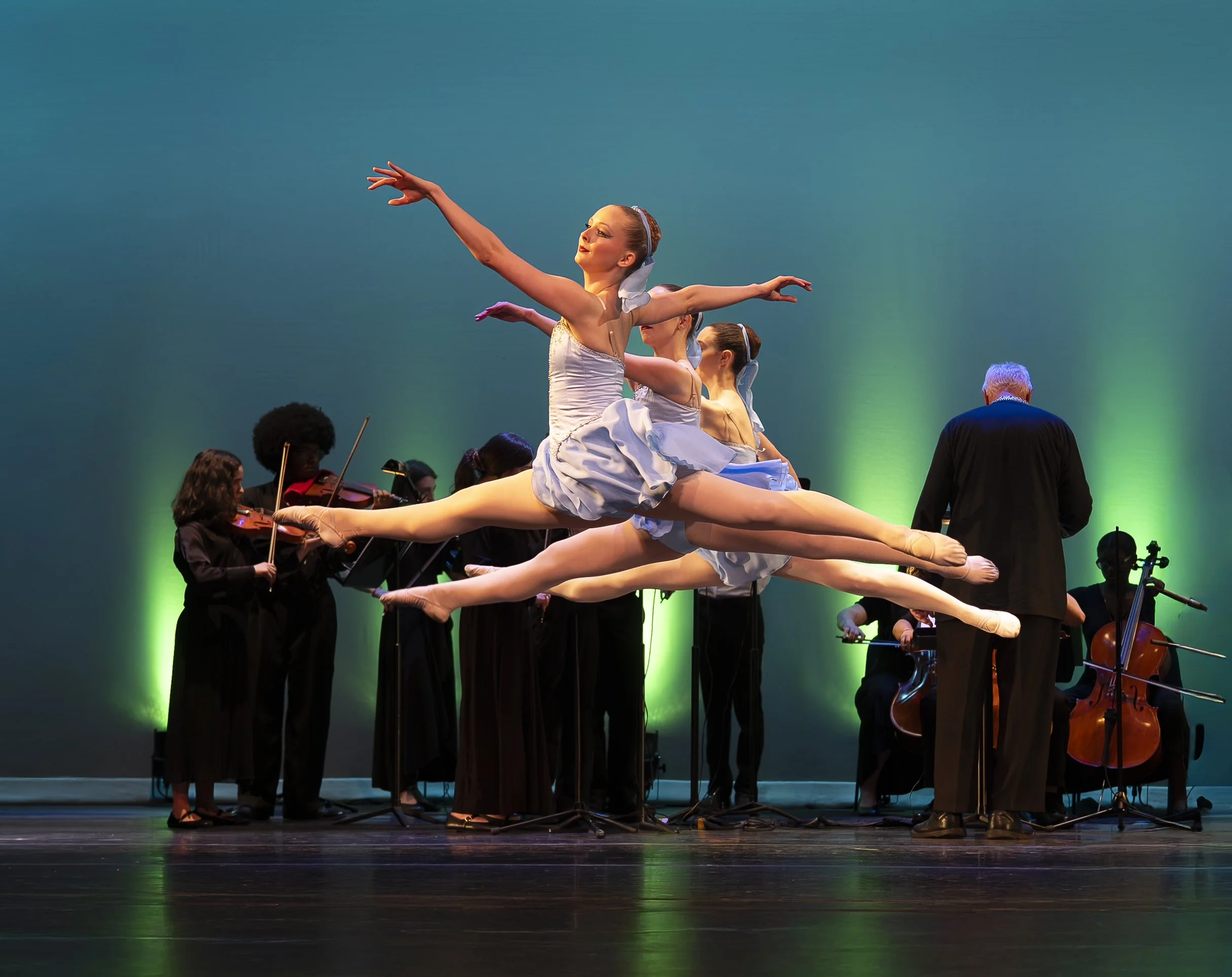 Ballet dancers perform a split on stage while musicians play string instruments in the background, illuminated by blue and green stage lighting.