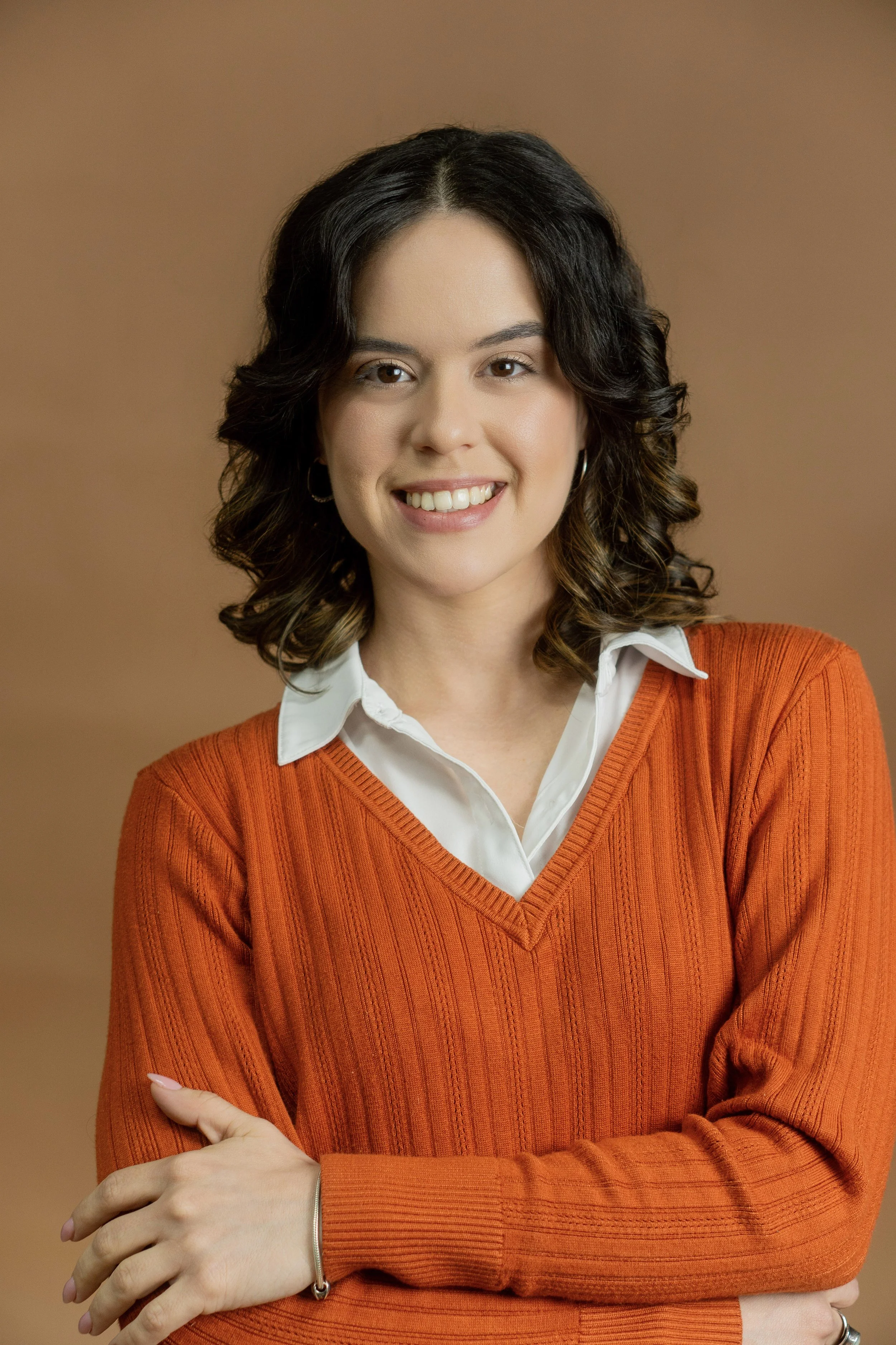 A woman with shoulder-length dark hair wearing a white shirt and an orange sweater, smiling with her arms crossed in front of a brown background.