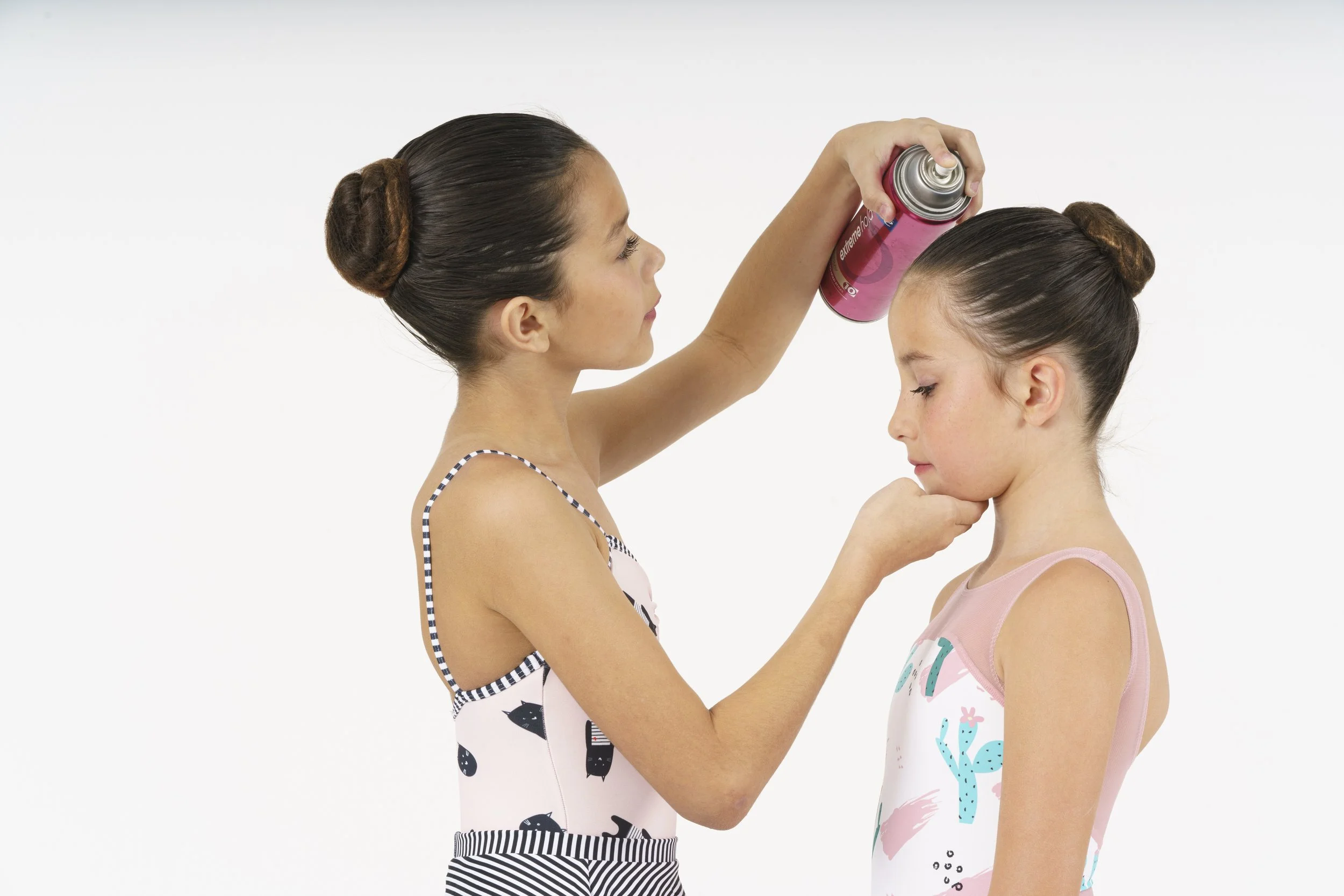 A woman spray paints the head of a young girl with brown hair tied in a bun, against a white background.